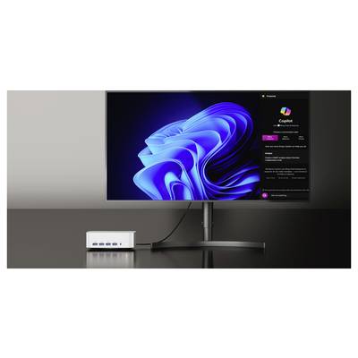 Ein Desktop-Monitor zeigt die Windows 11 Copilot-Oberfläche neben dem blauen Windows-Hintergrundbild an, mit einem kleinen Docking-Hub auf dem Schreibtisch darunter.