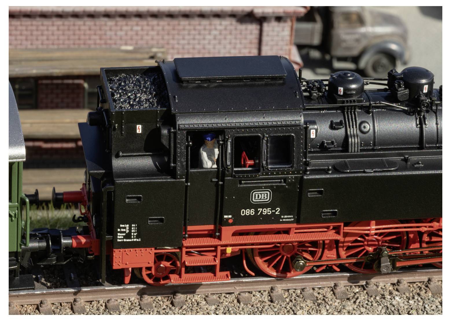 Schwarze Modelldampflokomotive auf Gleisen mit roten Rädern und einem sichtbaren Lokführer in der Kabine, fotografiert in Nahaufnahme vor einem unscharfen Backsteinzielhintergrund.