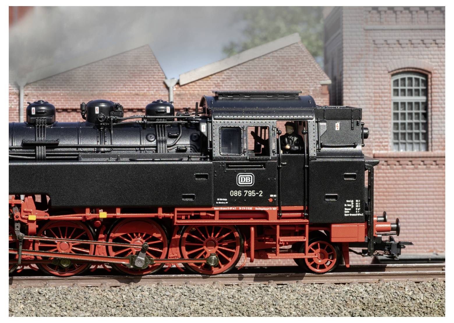 Schwarze Modell-Dampflokomotive mit der Nummer 086 795-2 auf Gleisen vor Ziegelbäuden, mit roten Rädern und Lokführer sichtbar in der Kabine.