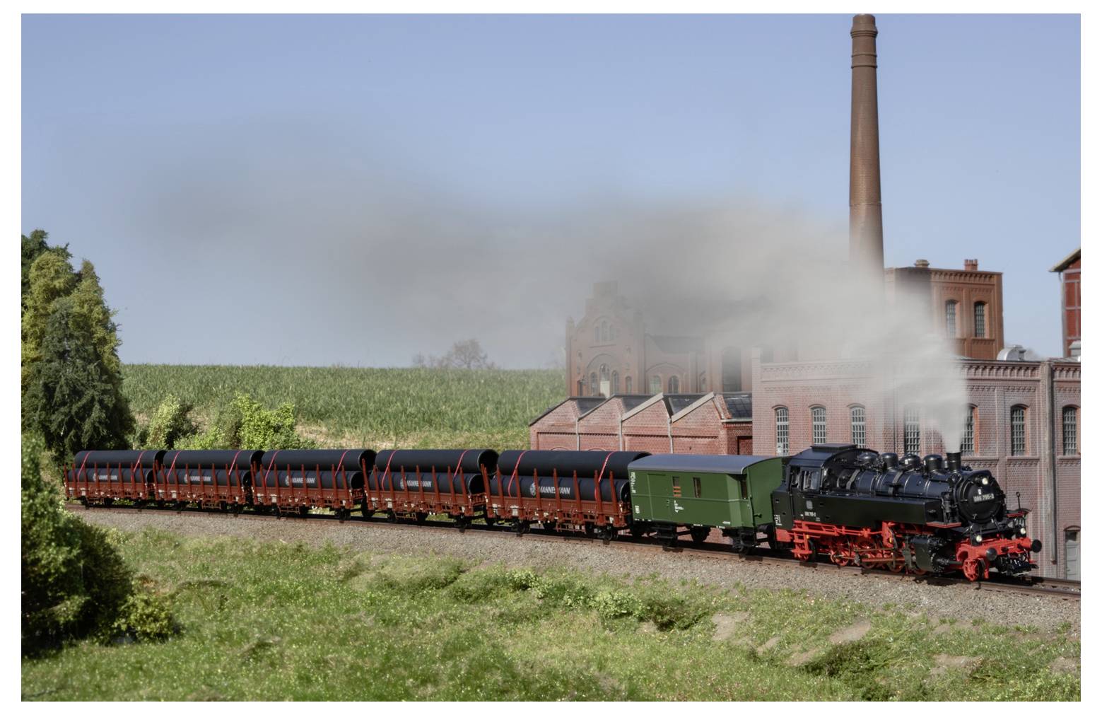 Dampflokomotive, die einen Güterzug durch ein ländliches Gebiet zieht, mit Rauch, der über ein industrielles Ziegelgebäude und einen hohen Schornstein im Hintergrund treibt.