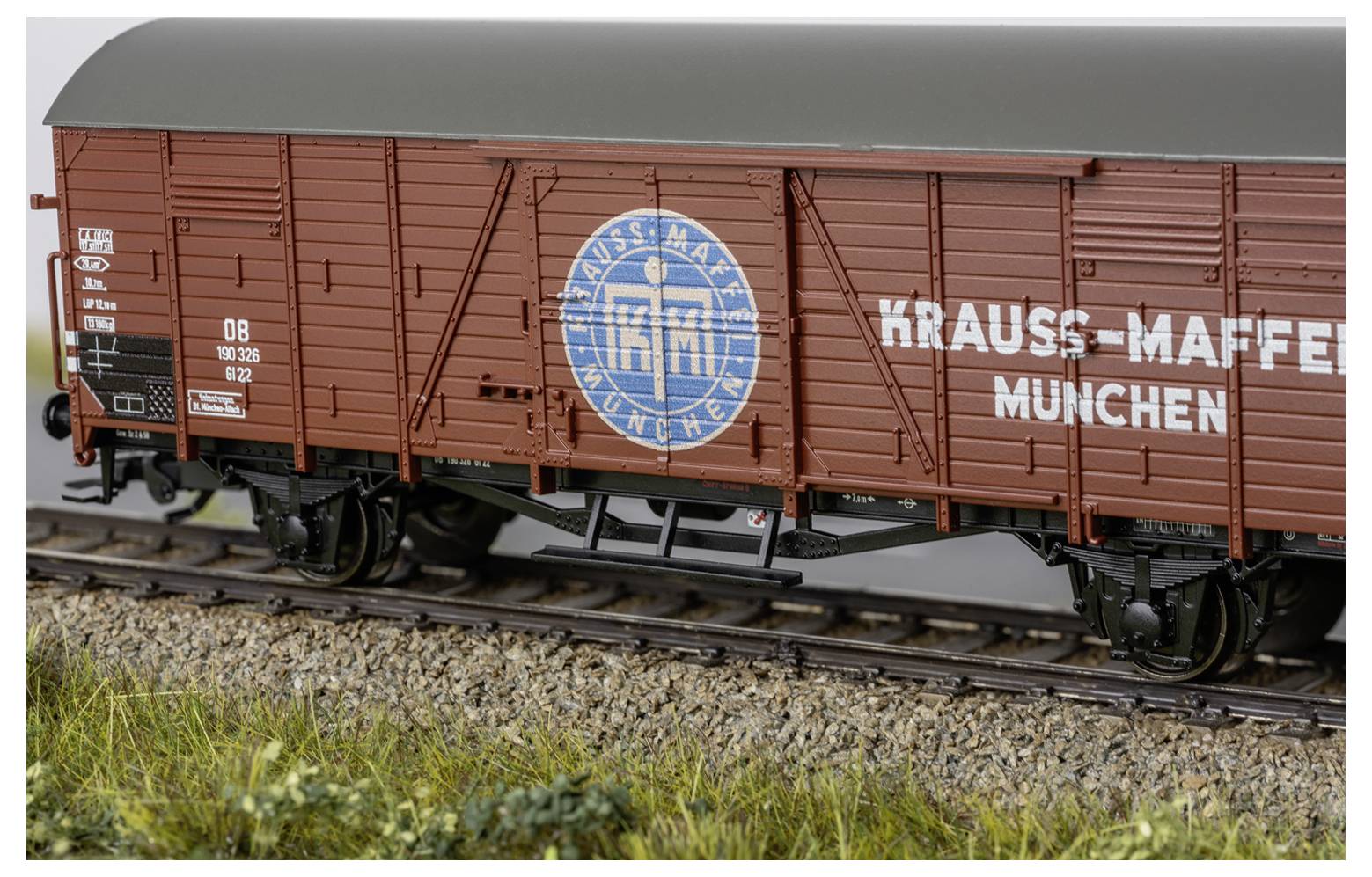 Modellgüterwagen auf Gleisen, braun mit "KRAUSS-MAFFEI MÜNCHEN" und einem blauen Kreislogo, dargestellt in einer grasigen Außenszene.