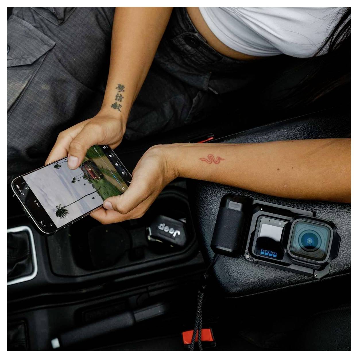 Person im Auto, der ein Smartphone hält, das eine Aussicht auf eine Küstenstraße zeigt, neben einer auf der mittleren Konsole montierten Action-Kamera.