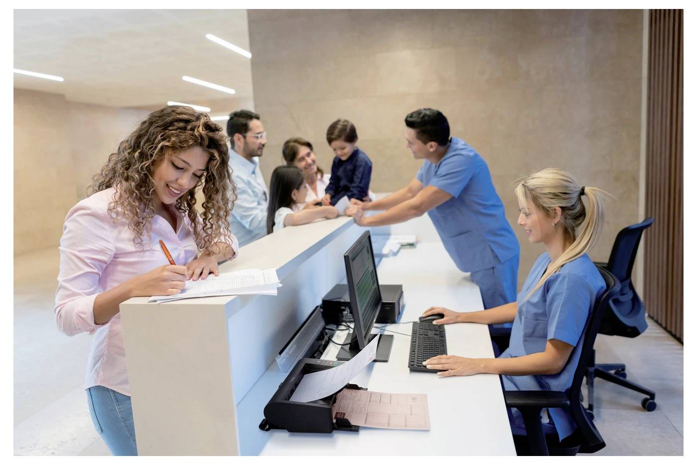 Krankenhaus-Empfangsbereich mit einer lächelnden Rezeptionistin am Computer, während eine Frau Formulare ausfüllt und eine Familie sich am Schalter anmeldet.