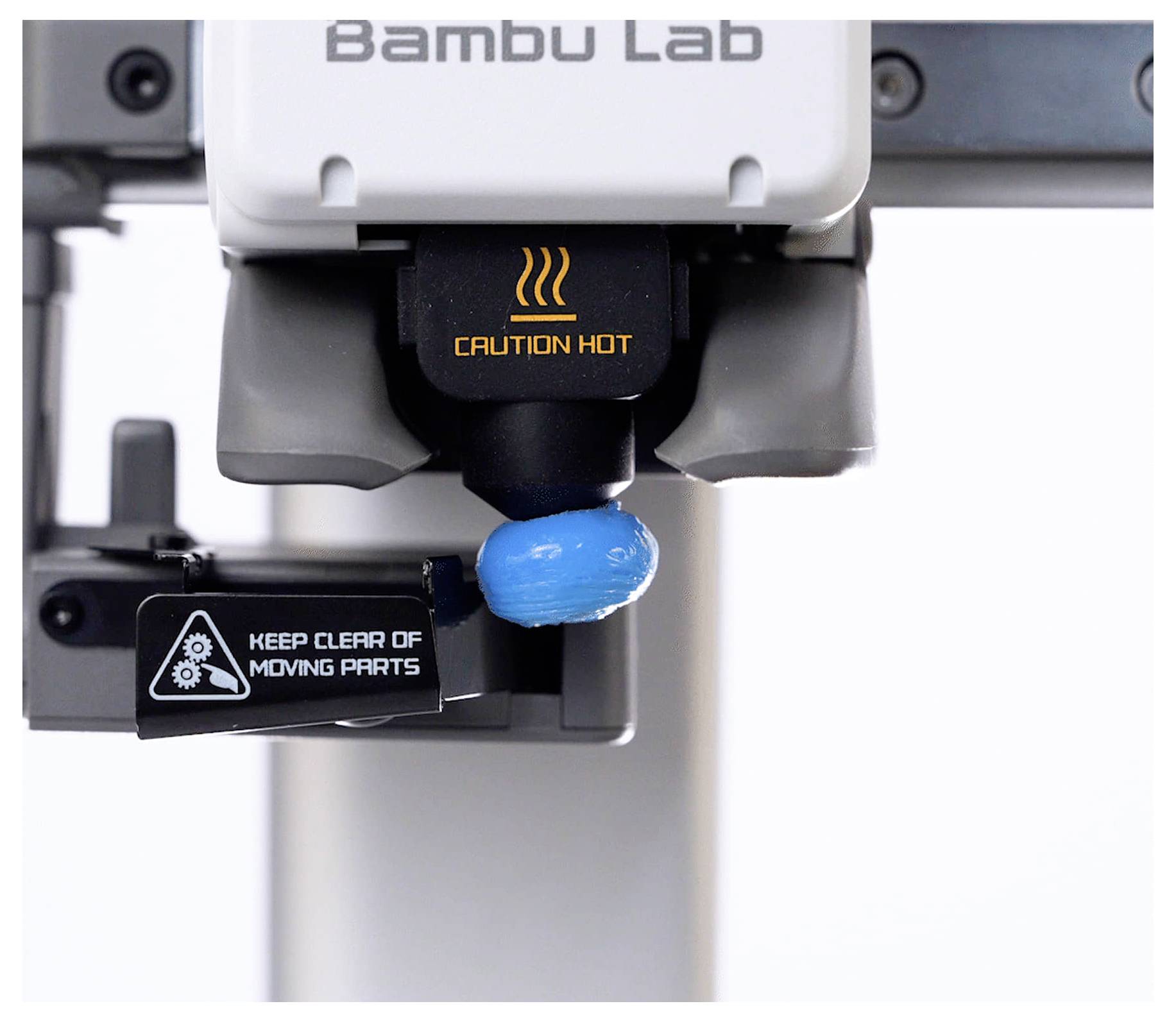 Nahaufnahme eines Bambu Lab 3D-Druckers-Extruders beim Drucken eines blauen Objekts, wobei die Warnhinweise „CAUTION HOT" und „KEEP CLEAR OF MOVING PARTS" sichtbar sind.
