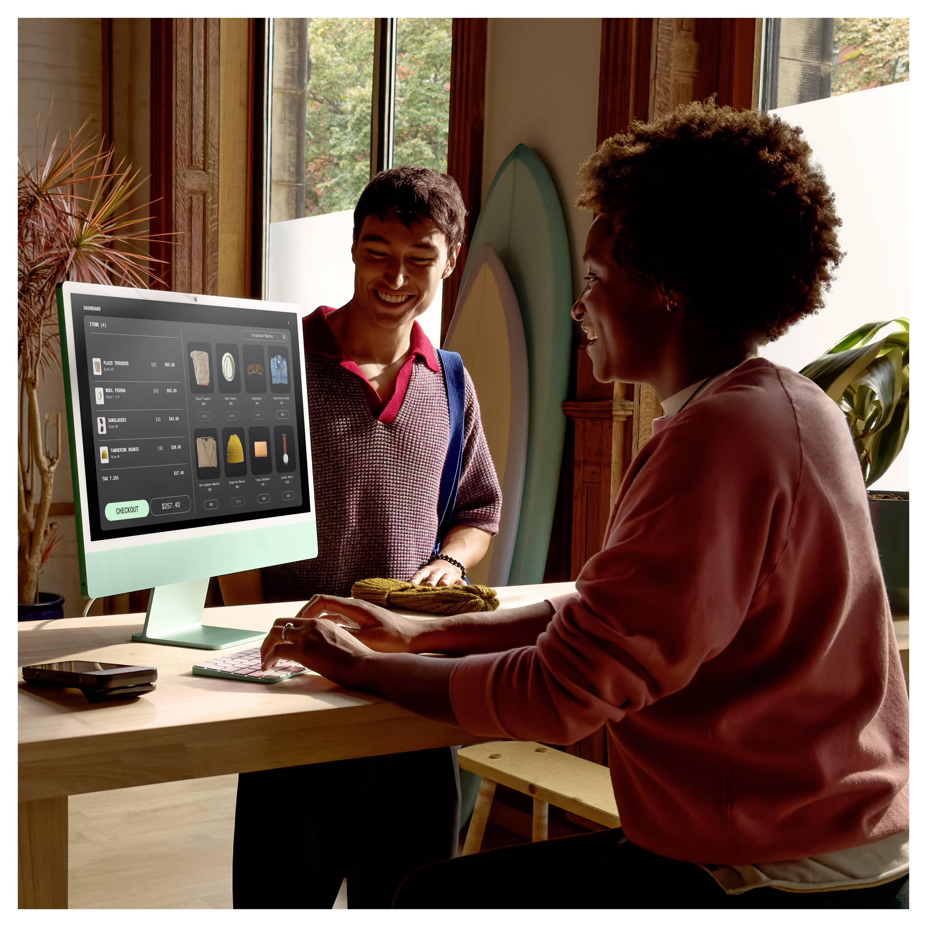 Zwei Personen an einem Desktop-Computer in einem hellen Raum, eine steht und lächelt, während die andere an einer kassenähnlichen Schnittstelle auf dem Bildschirm tippt.
