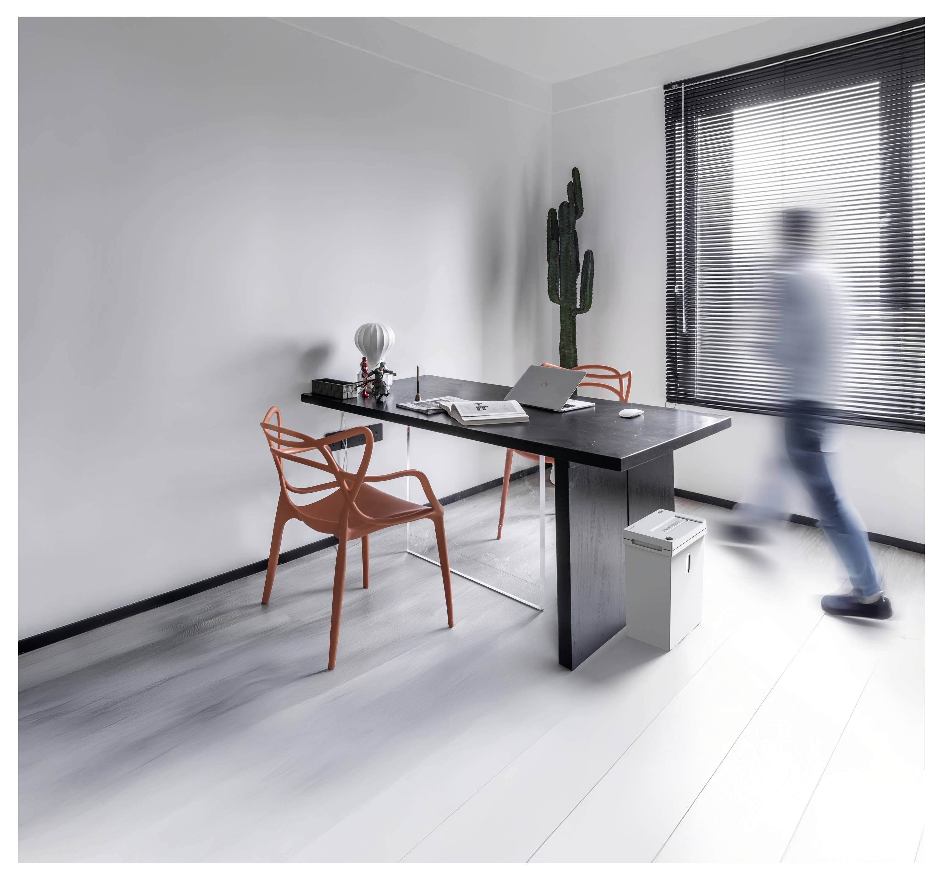 Minimalistische moderne Büroraum mit schwarzem Schreibtisch, orangefarbenen Stühlen und einer unscharfen Person, die an einem Fenster mit Jalousien vorbeigeben.