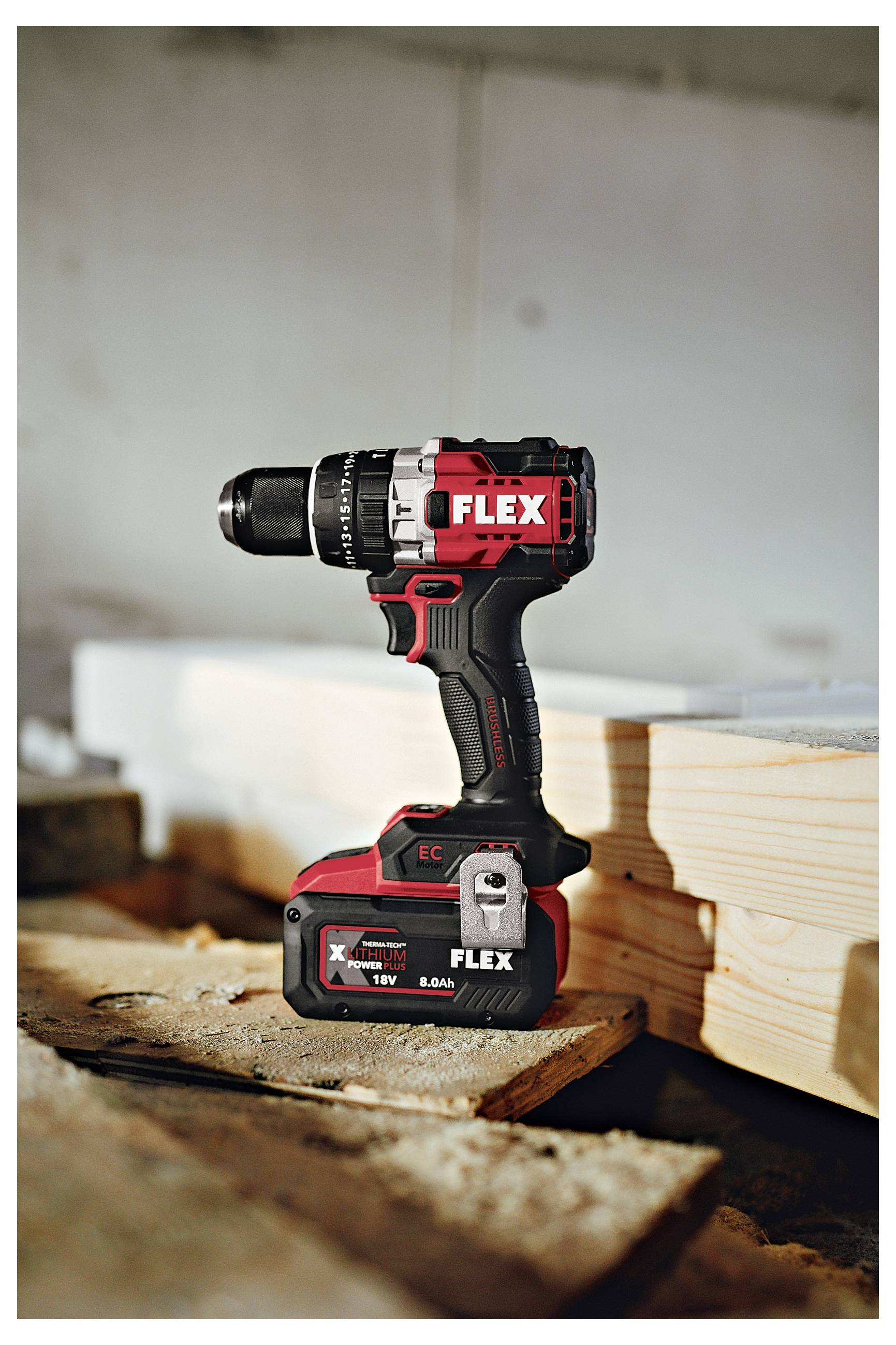 Rote und schwarze FLEX-Akku-Bohrmaschine, die auf einer Holzwerkbank neben gestapeltem Bauholz in einer Werkstatt steht.