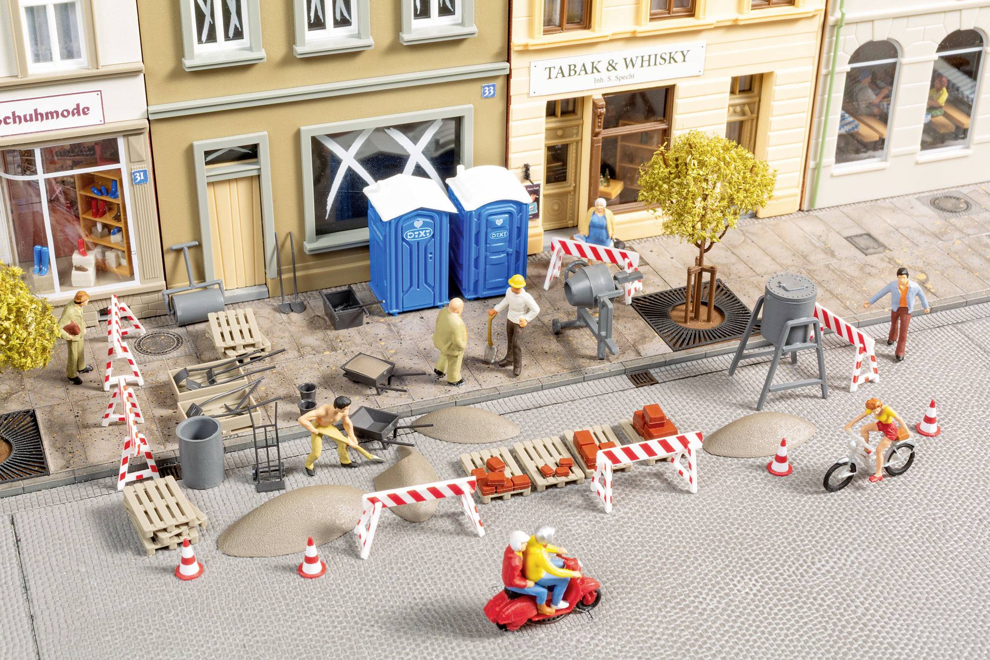 Miniatur-Straßenbauszene vor Geschäften mit Straßensperren, Arbeitern, Maschinen, tragbaren Toiletten und vorbeigehenden Personen.