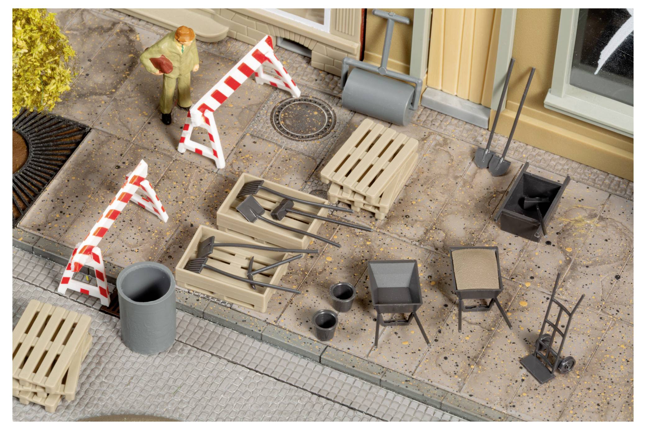 Miniatur-Straßenbauszene mit Absperrungen, Werkzeugen, Paletten, Behältern und einer Arbeiterfigur auf einem Gehweg