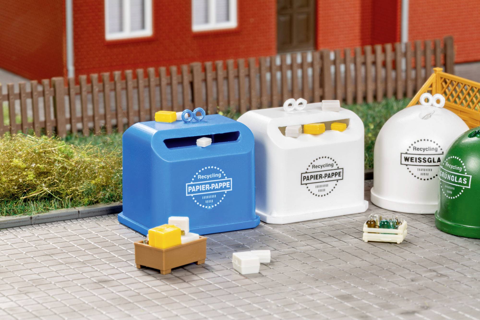 Miniatur-Recyclingbehälter für Papier, Glas und Mischabfälle aufgereiht im Freien neben einem Zaun und einer Hauswand.