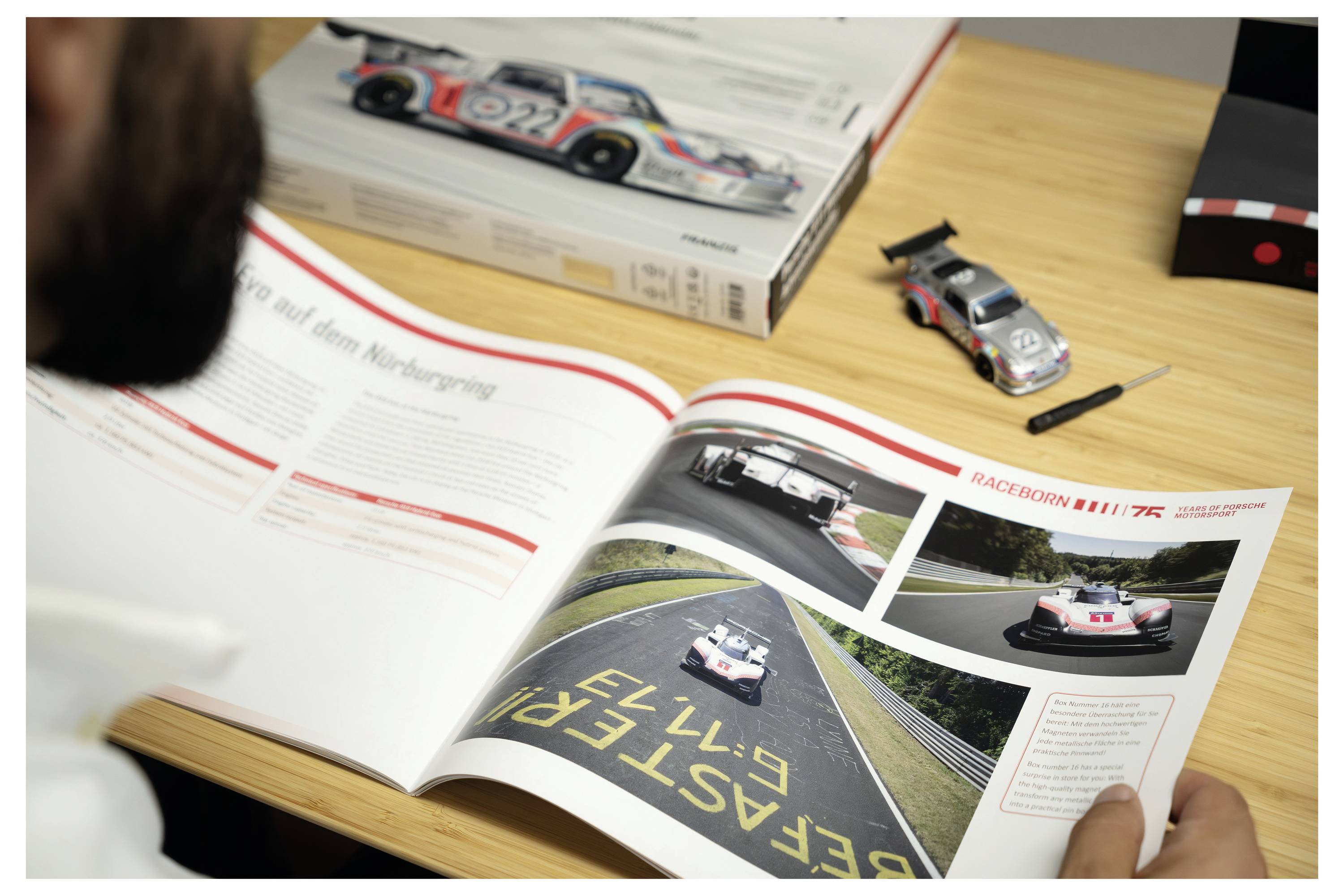 Person liest eine Rennfahrzeugzeitschrift mit Fotos eines Porsche-Rennwagens auf einem Holztisch, neben einem Spielzeugauto und gestapelten Modellautokästen.