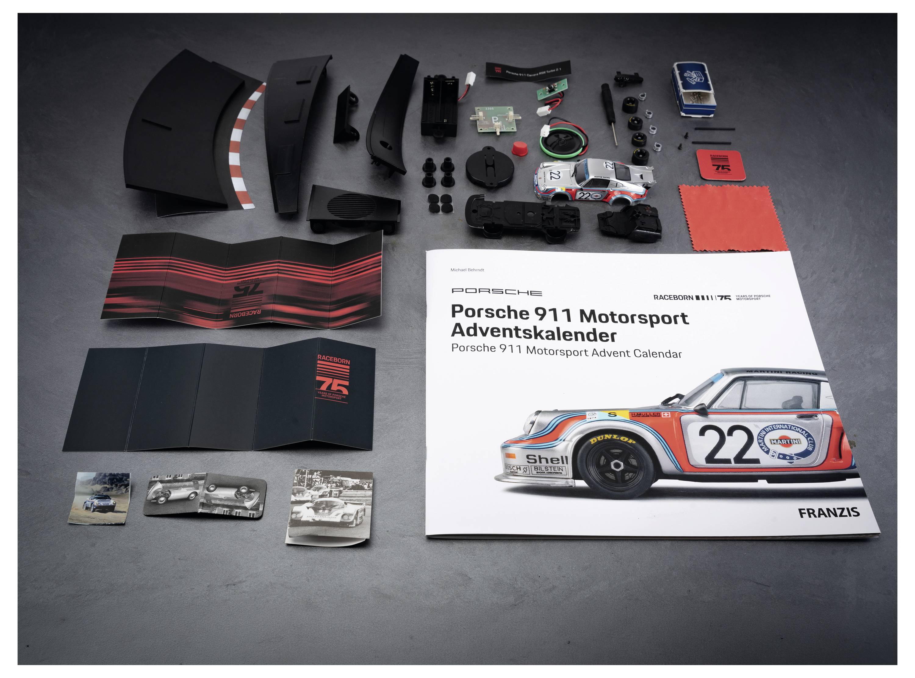 Porsche 911 Motorsport Adventskalender Geschenkset mit Autoteilen, Zubehör, Anleitungsheft und Kalenderkomponenten, angeordnet auf einer grauen Oberfläche.
