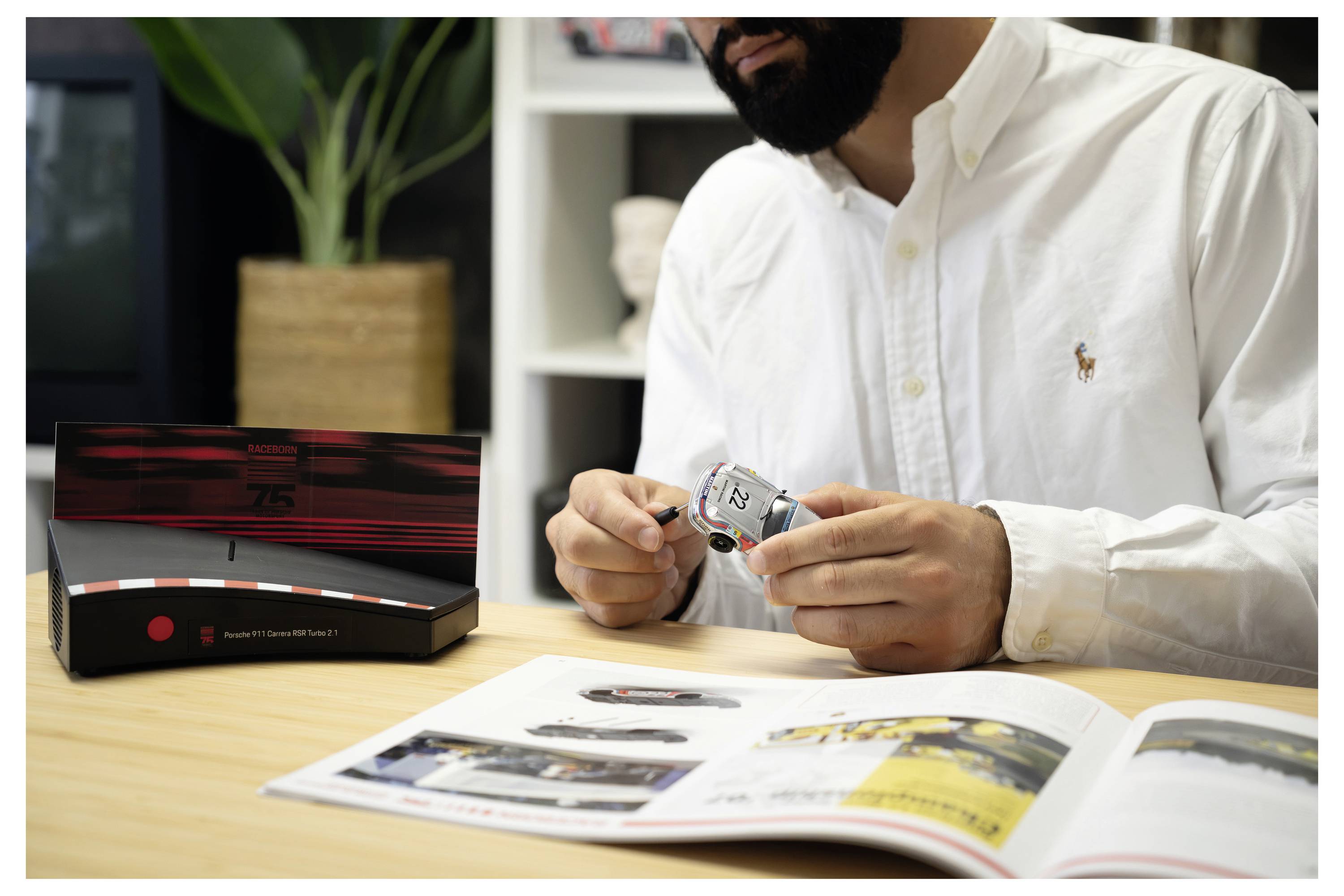 Eine Person hält eine Porsche 911 Motorsport Adventskalender-Box in einem Wohnzimmer, mit Regalen und einem Stuhl im Hintergrund.