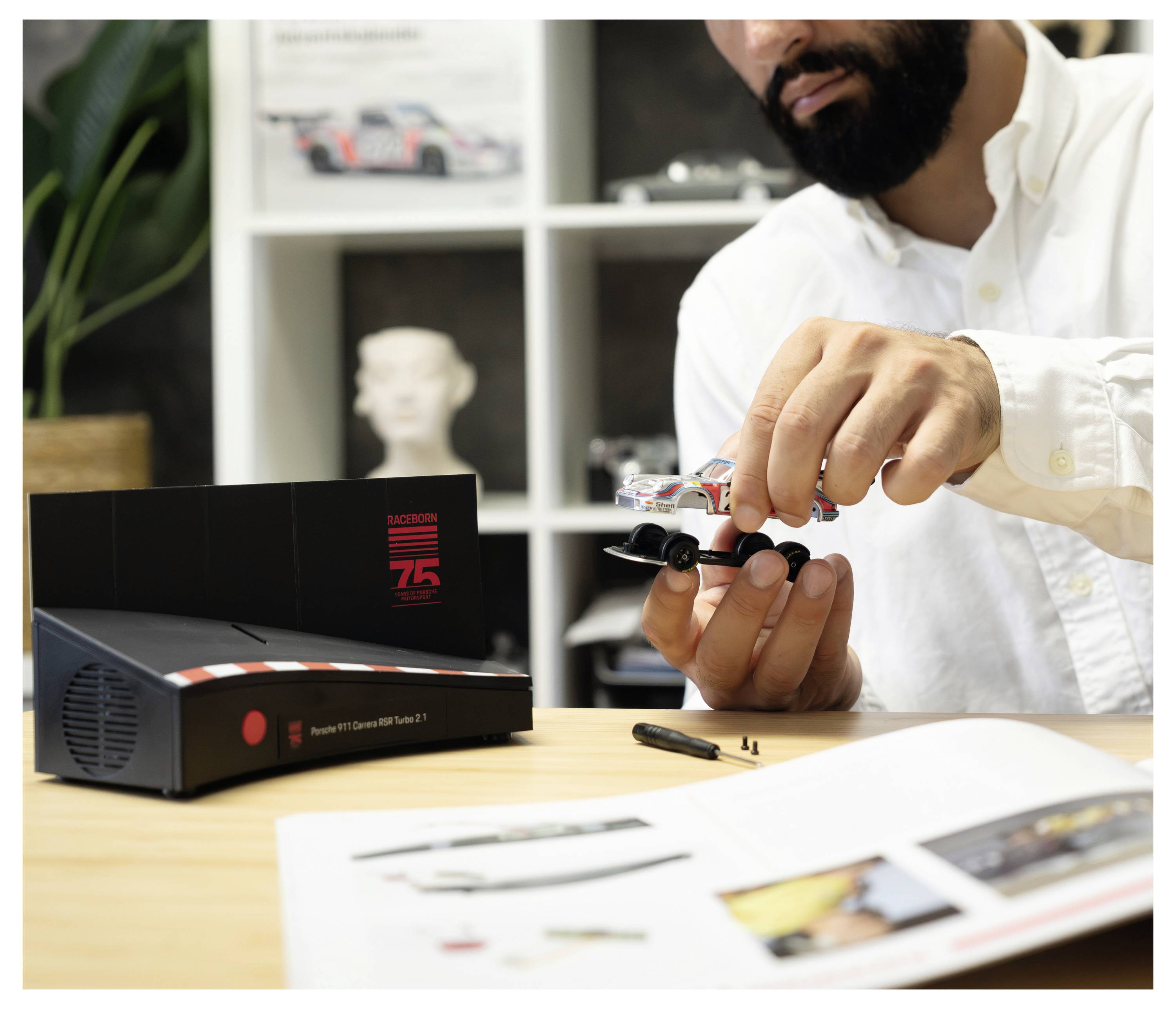 Ein Mann montiert ein kleines Modellauto auf einem Schreibtisch neben einer offenen Elektronikbox und einer Bedienungsanleitung.