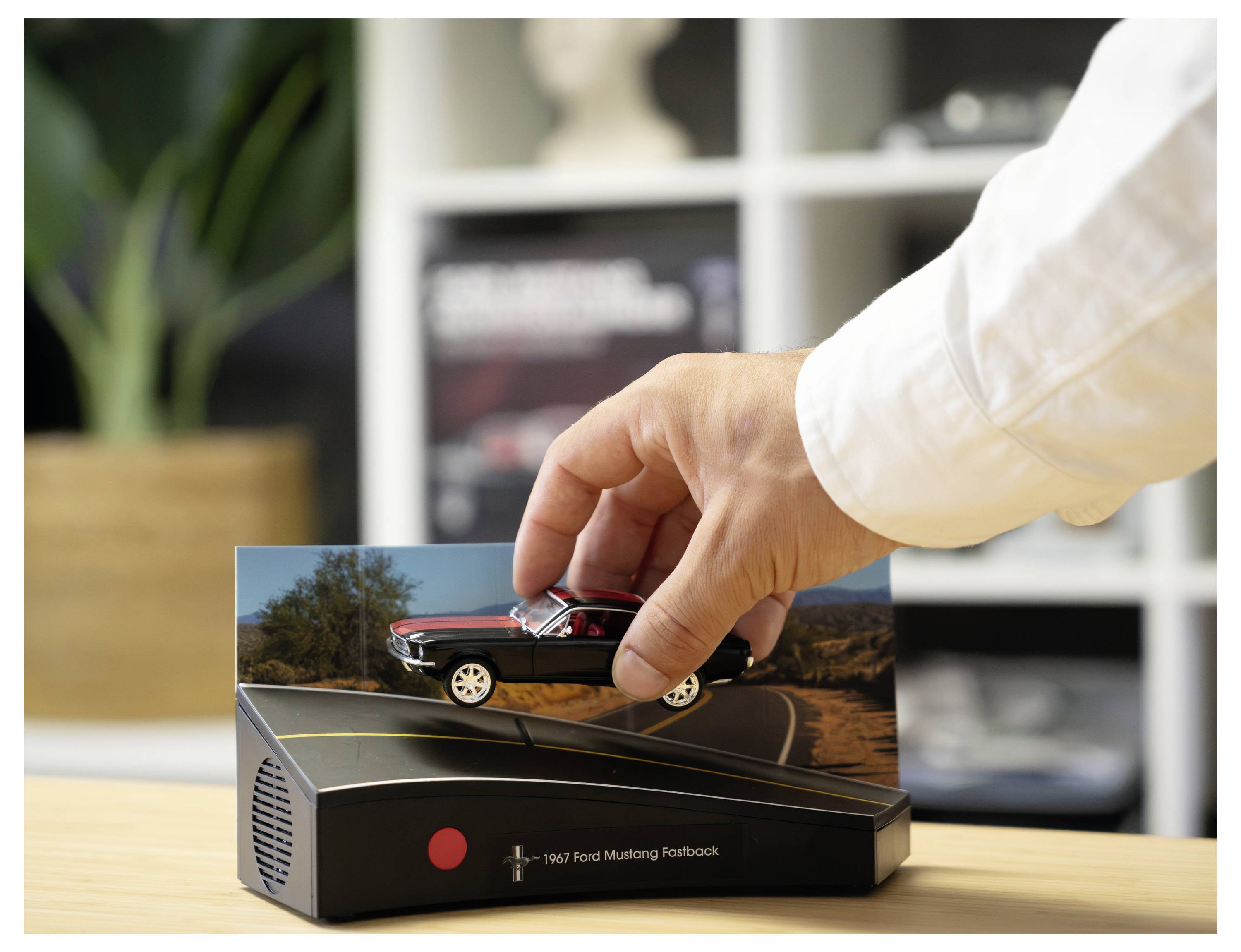 Hand, die einen rot-schwarzen Spielzeug-Ford Mustang auf einen Ausstellungssockel mit der Aufschrift „1967 Ford Mustang Fastback" setzt.