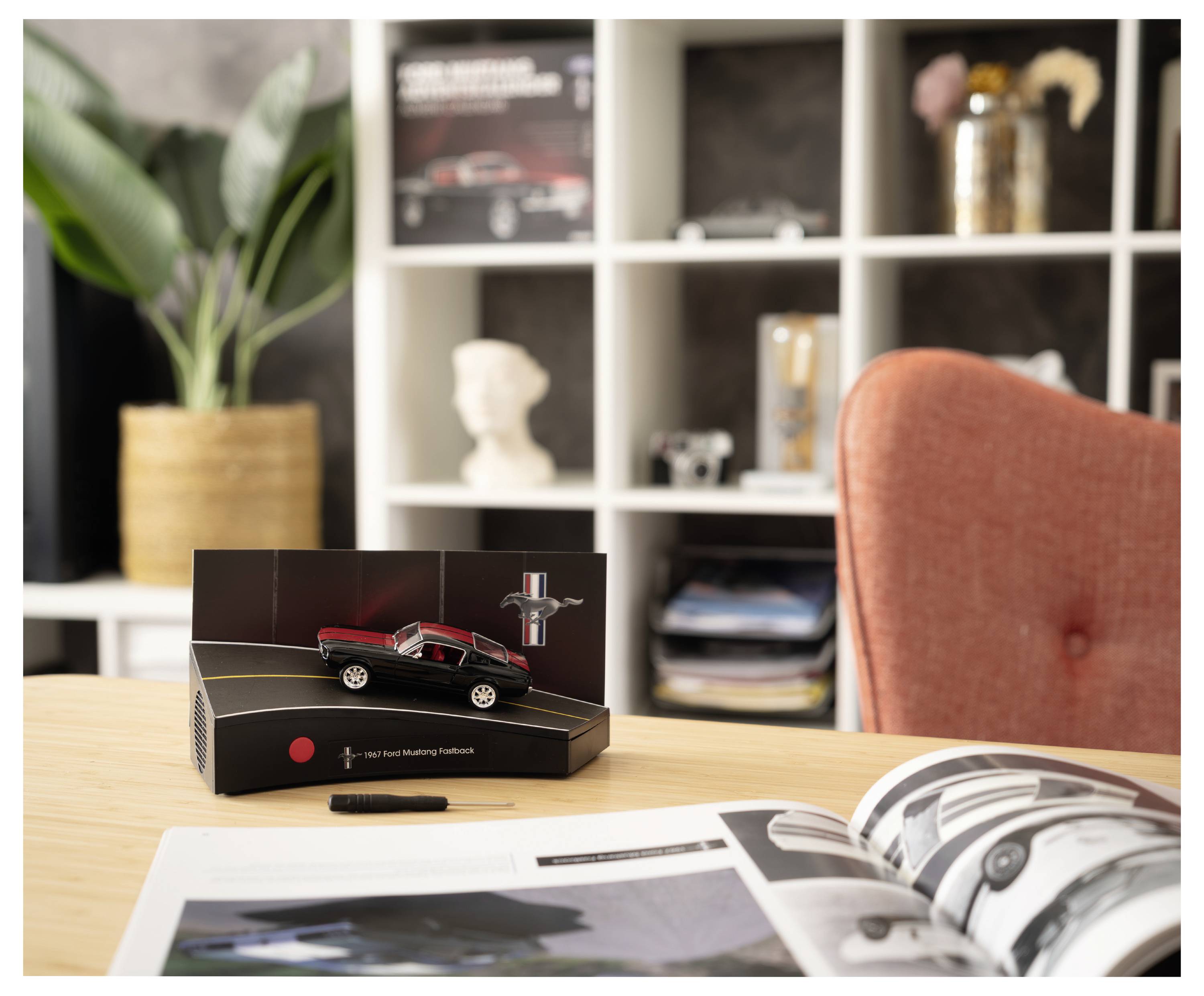 Ein rot-schwarzes Modellauto, das auf einer Tischbox ausgestellt ist, mit einer offenen Zeitschrift im Vordergrund und einem Homeoffice-Regal im Hintergrund.