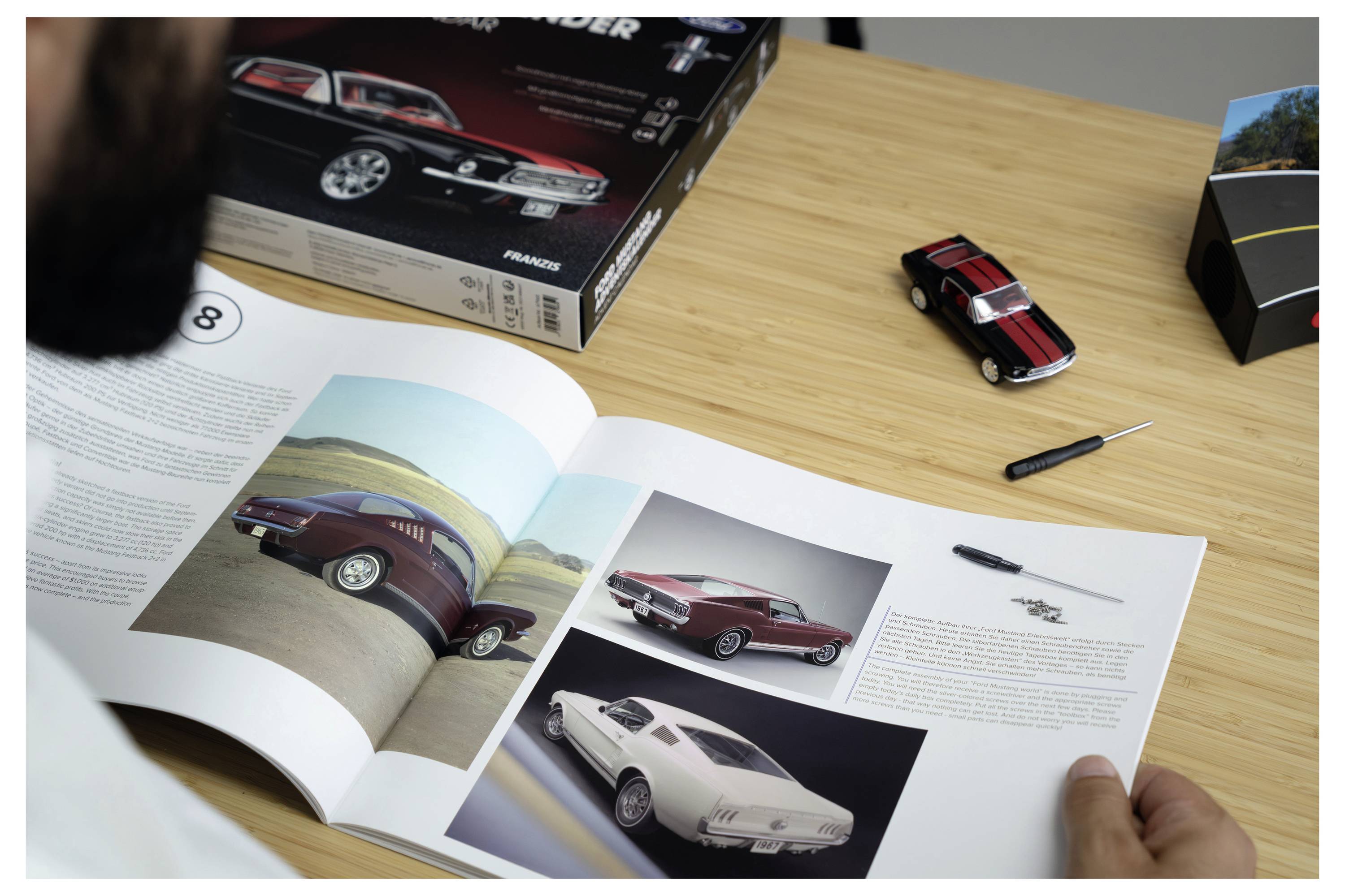 Eine Person liest ein offenes Buch über Autodesign an einem Schreibtisch, mit einem roten klassischen Automodell und einem verpackten Autobausatz in der Nähe.