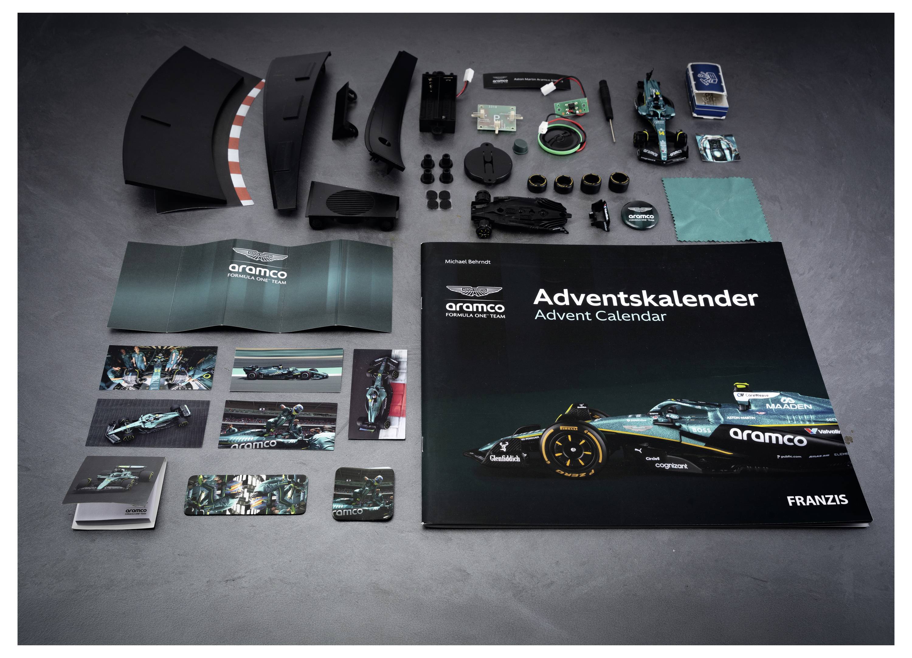 Unzusammengesetztes Aramco Adventskalender-Set mit schwarzer Schachtel, nummerierten Teilen, Mini-Autoteilen, Karten und ausklappbarem Booklet auf einer grauen Oberfläche ausgelegt.