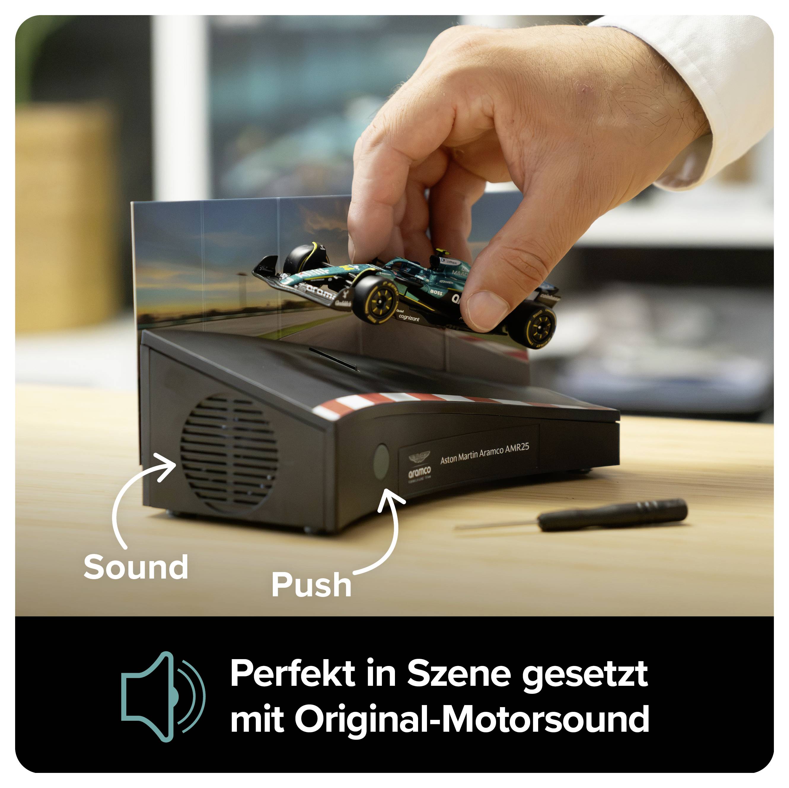 Hand platziert ein Formel-1-Modellauto auf einem schwarzen Display mit Lautsprecher; Text: 'Perfekt in Szene gesetzt mit Original-Motorsound'.