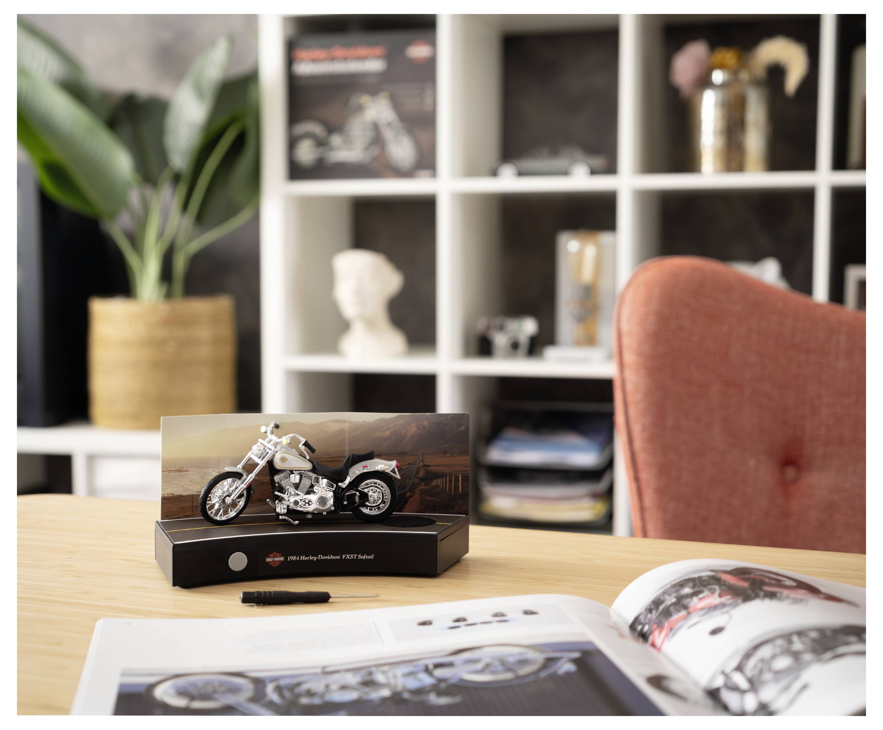 Miniatur-Motorradmodell auf einem Ausstellungsständer auf einem Holztisch, mit einer offenen Zeitschrift und einem roten Stuhl im Hintergrund eines Homeoffice.