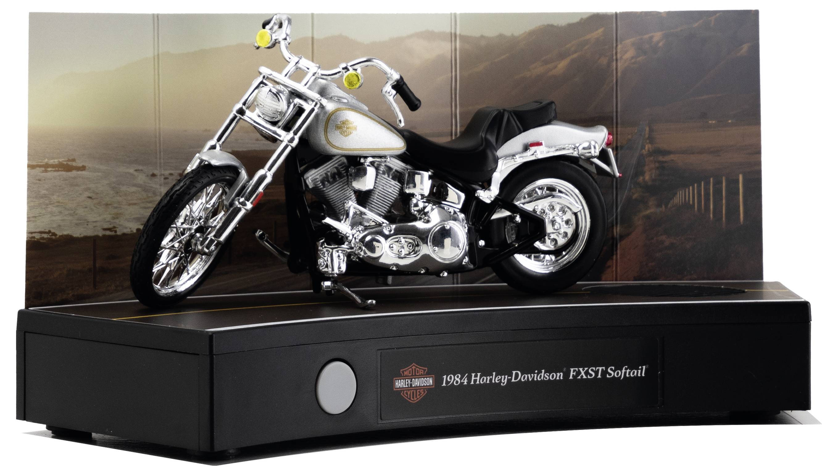 Modellmotorrad-Ausstellung eines 1984er Harley-Davidson FXST Softail auf schwarzem Sockel, dargestellt in Chrom und Weiß mit landschaftlichem Hintergrund