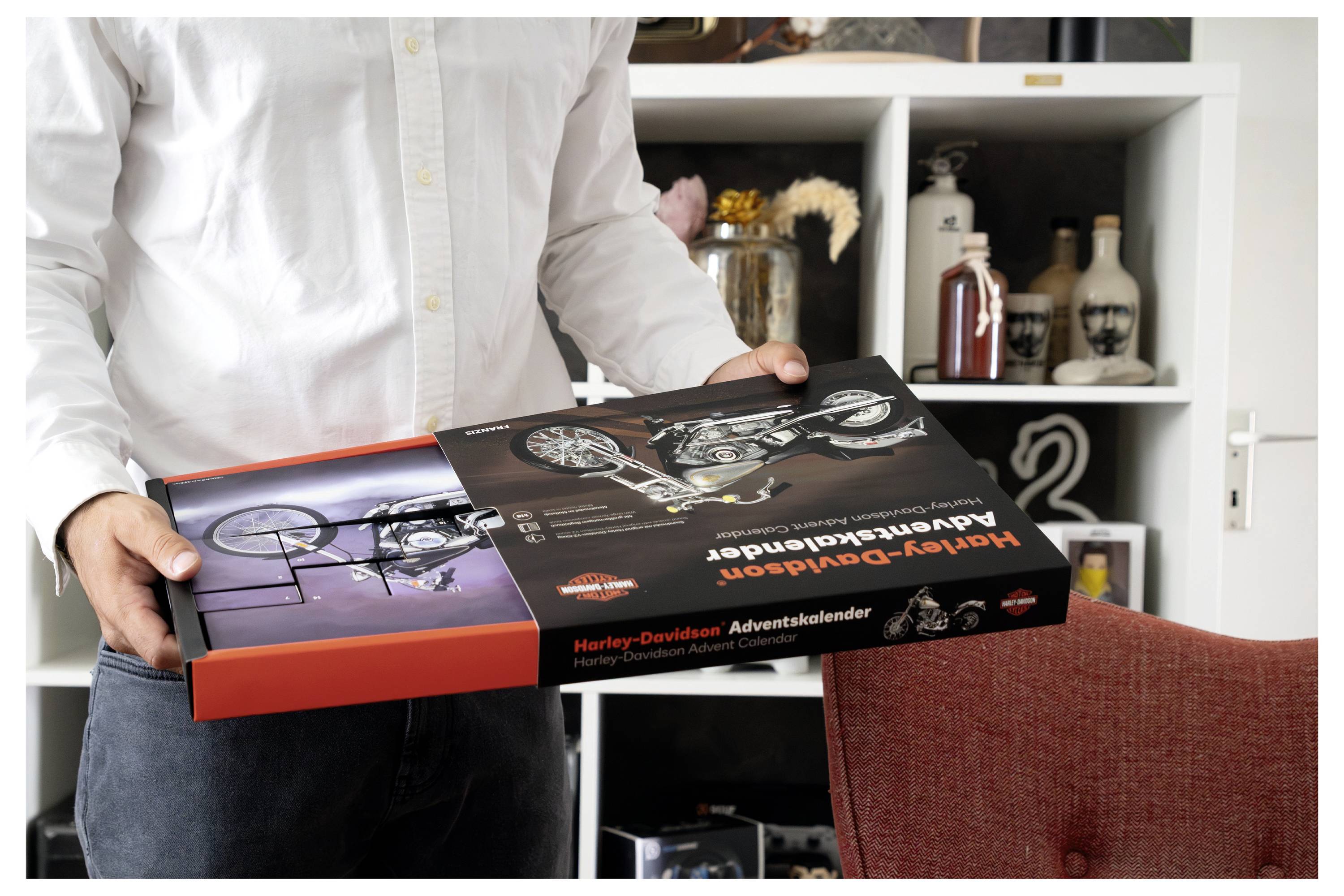 Eine Person hält eine Harley-Davidson Adventskalender-Box in einem Wohnzimmer, die motorrad-thematische Verpackung und Bilder auf dem geöffneten Deckel zeigt.