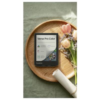 PocketBook Verse Pro Color E-Reader auf einem Holztablett neben Blumen und einer aufgerollten Serviette auf grünem Stoffhintergrund.