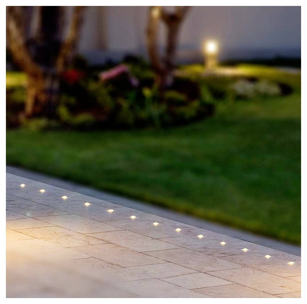 Terrasse im Freien mit eingelassenen Bodenleuchten entlang der Kante, neben einem unscharfen Rasen und Garten in der Dämmerung.