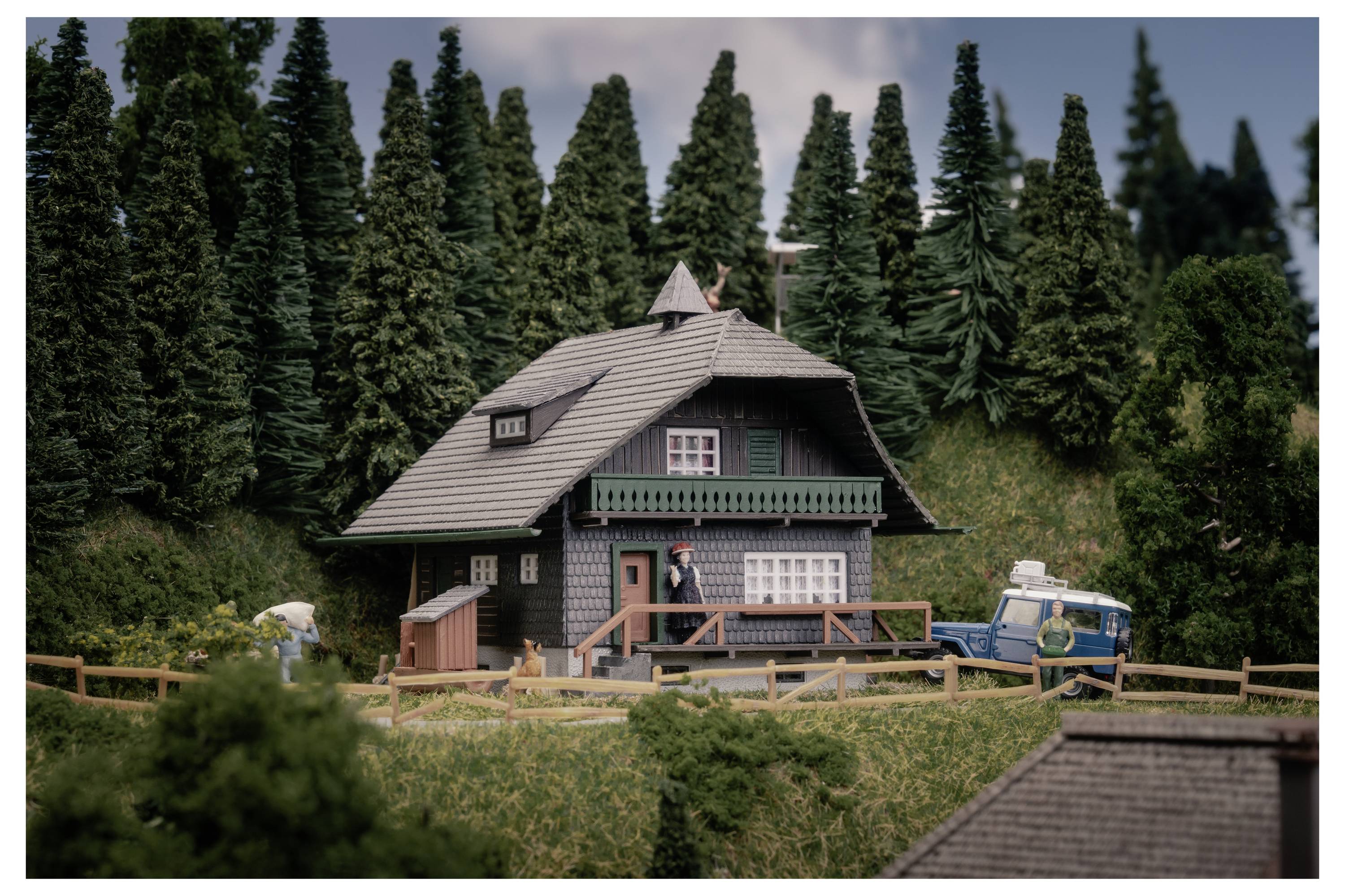 Miniatur-Landhaus-Modell mit einer Person draußen, umgeben von dichten Nadelbäumen und einem blauen Fahrzeug, wie eine kleine Dorfszene.