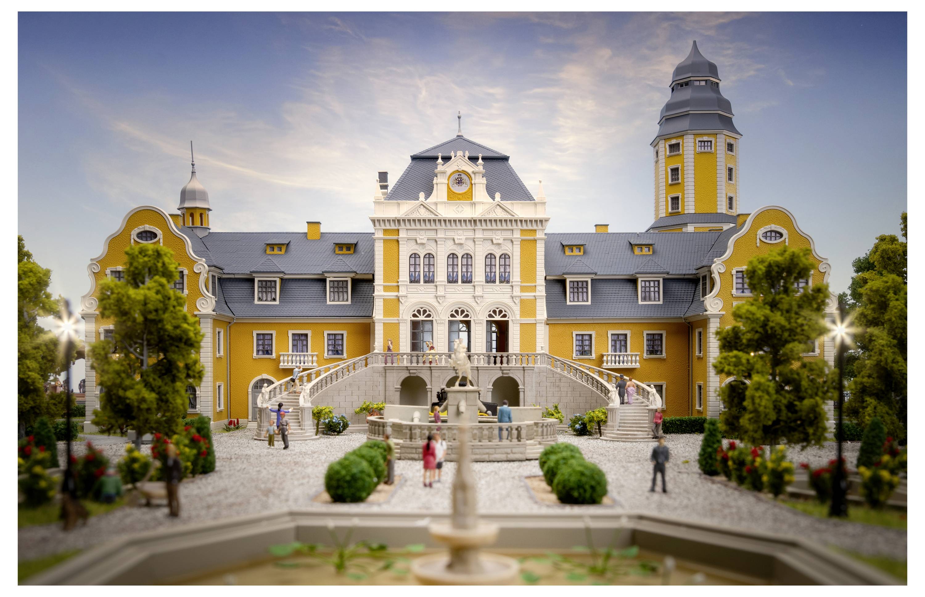 Ein Modell eines prächtigen gelb-weißen historischen Herrenhauses mit doppelten Türmen, zentraler Treppe und Brunnen davor, gezeigt in einem angelegten Innenhof.