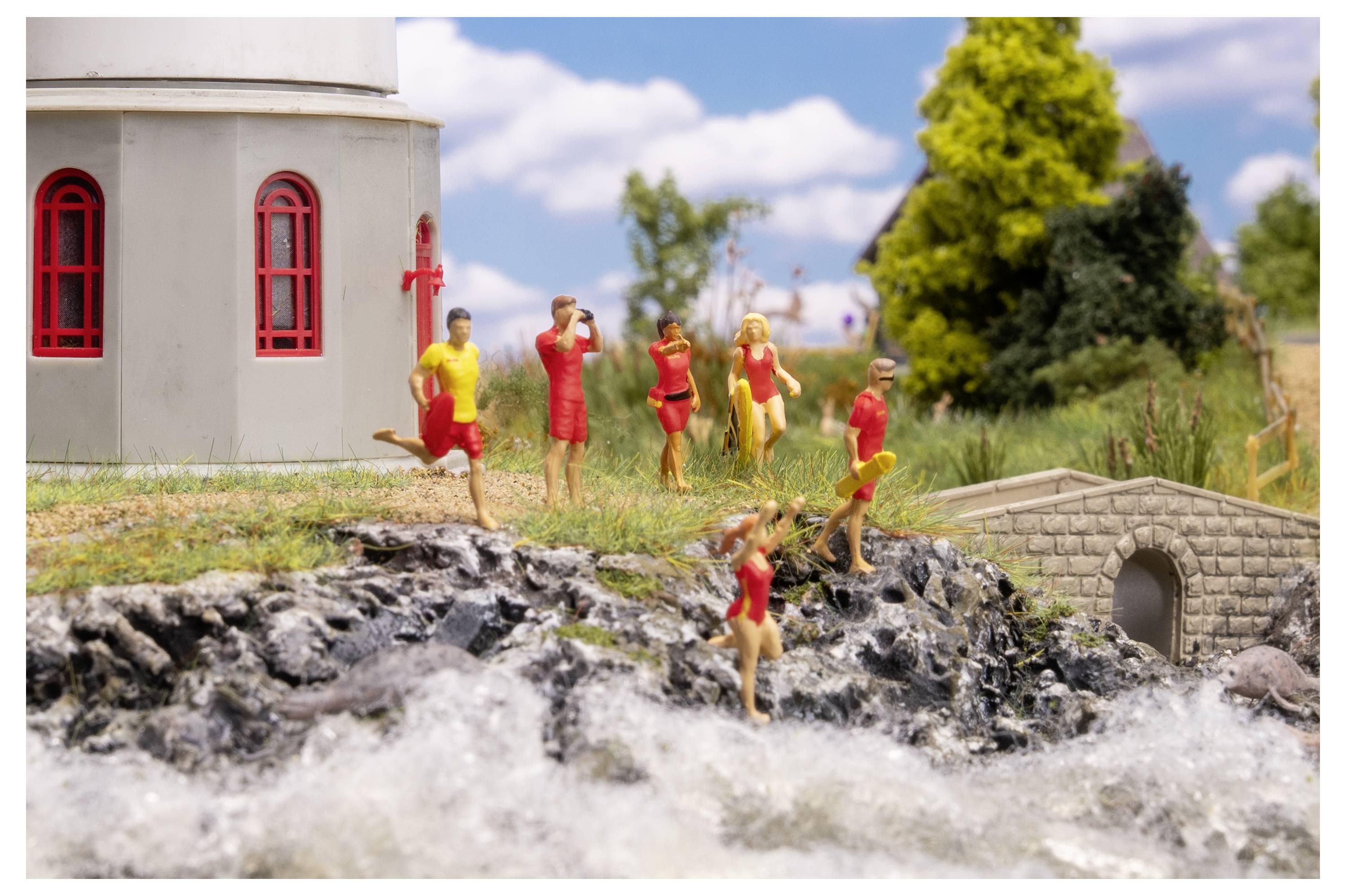 Miniaturszene mit Menschen in roter und gelber Kleidung, die auf felsigem Boden stehen und rennen neben einer kleinen Steinbrücke und einem kirchenähnlichen Gebäude in der Nähe von Wasser.