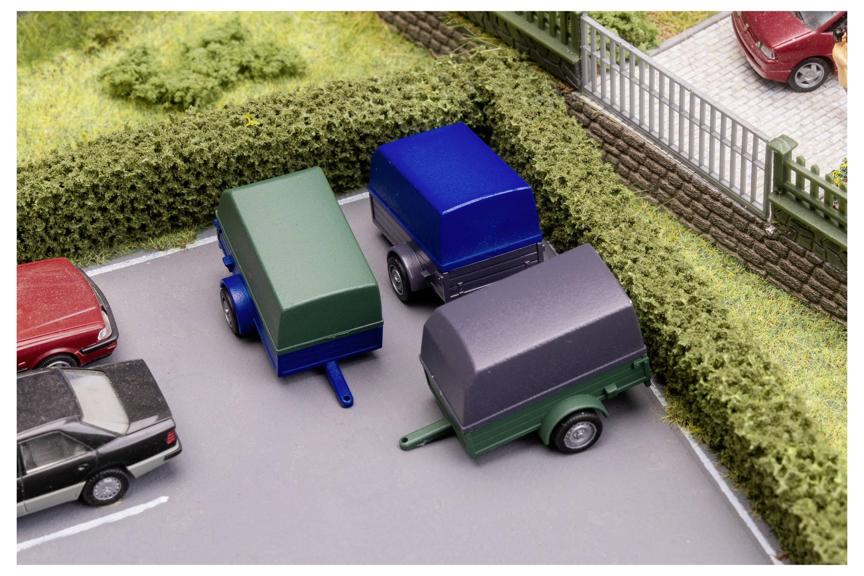 Drei abgedeckte Nutzanhänger, die auf einem Platz neben Hecken und einem Metallzaun geparkt sind, mit anderen Autos, die in der Nähe teilweise sichtbar sind.