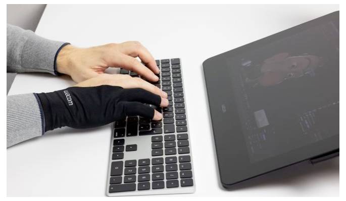 Zwei Personen tippen auf einer Laptop-Tastatur, eine trägt einen schwarzen Handschuh, neben einem Touchscreen-Tablet auf einem weißen Schreibtisch.