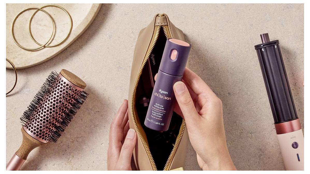 Hände platzieren eine Dyson Chitosan Haarserum-Flasche in eine offene Kosmetiktasche, mit einer Rundbürste, einem Haarstyling-Gerät und Haargummis in der Nähe.