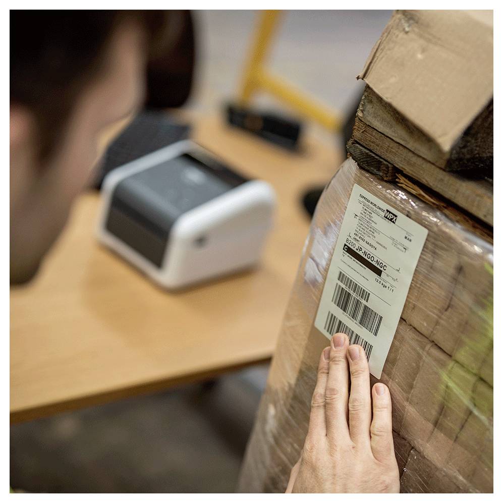 Ein Hand, die ein Versandetikett auf einem Kartonpaket auf einem Tisch scannt, mit einem unscharfen Etikettendrucker im Hintergrund.