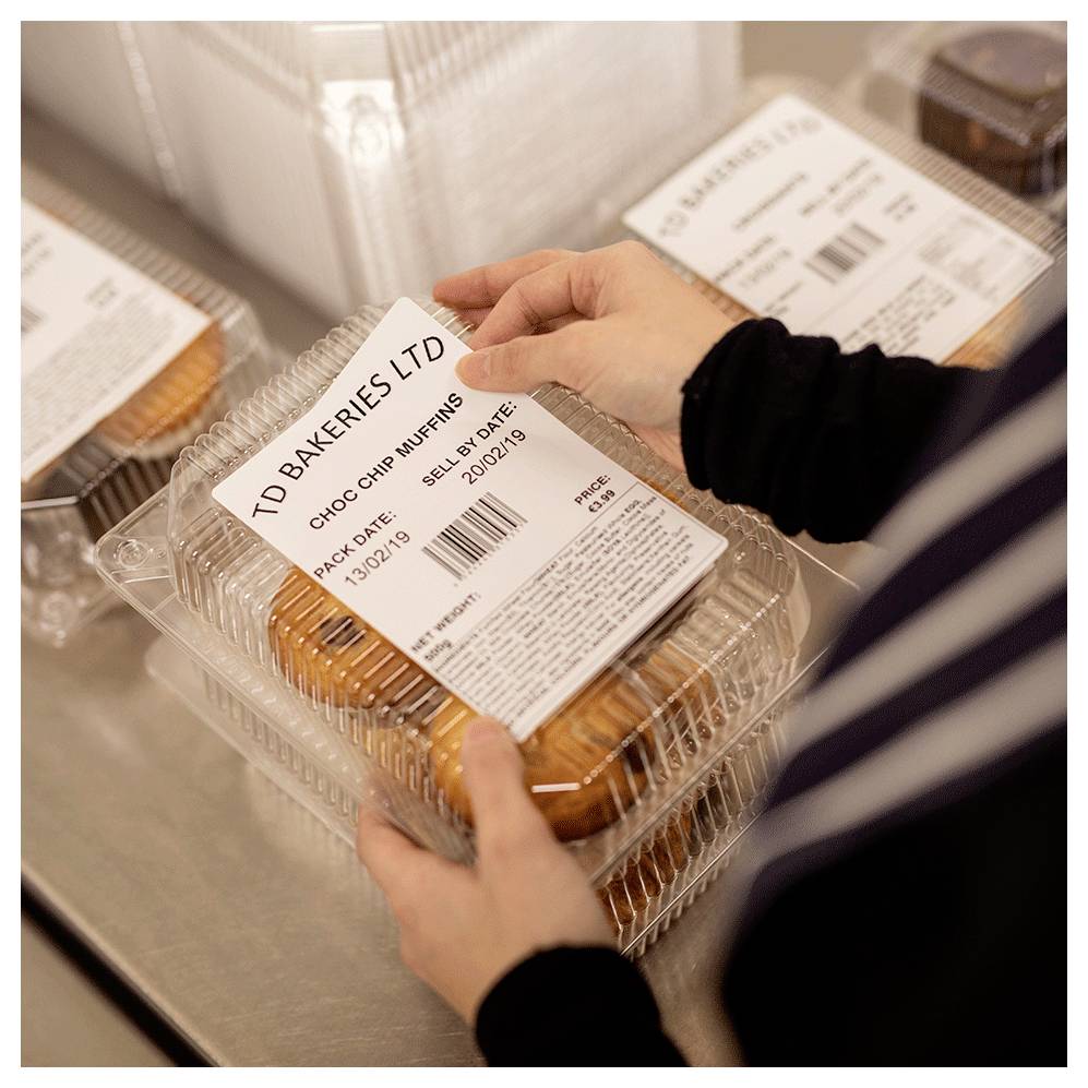 Hände, die eine Kunststoff-Klappschachtel mit "Choc Chip Muffins" mit der Bezeichnung "TD Bakeries Ltd" auf einem Förderband oder Packtisch ablegen.