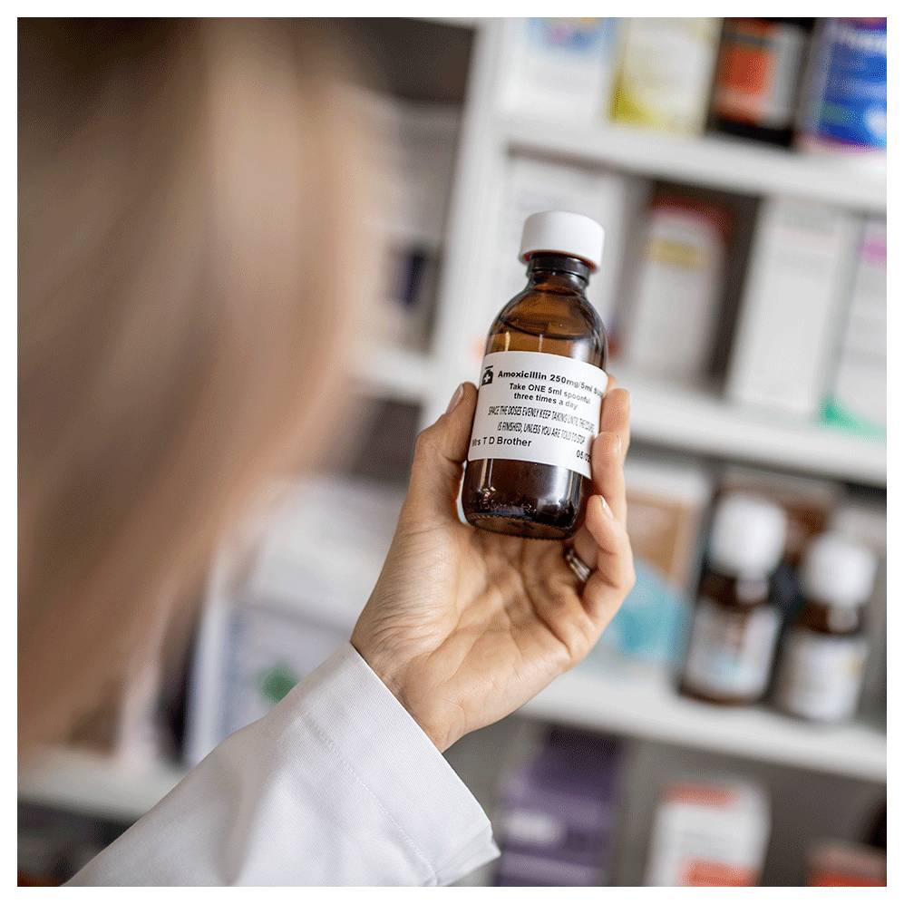 Eine Person in einem weißen Kittel hält eine bernsteinfarbene Arzneimittelflasche vor Apotheken­regalen.