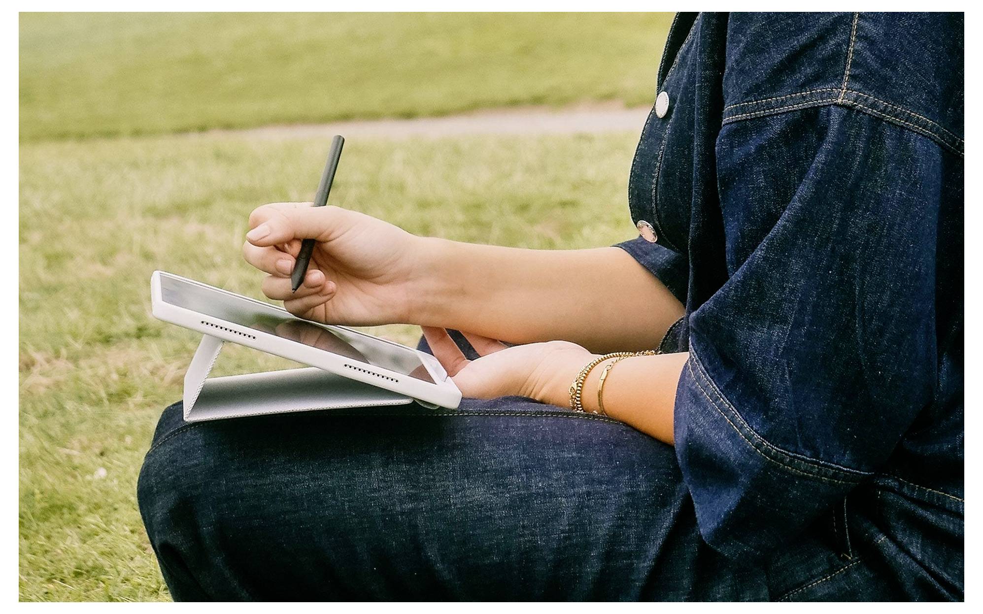 Person sitzt draußen auf Gras und benutzt einen Stylus auf einem kleinen Tablet oder Zeichengerät, das auf ihrem Schoß ruht.
