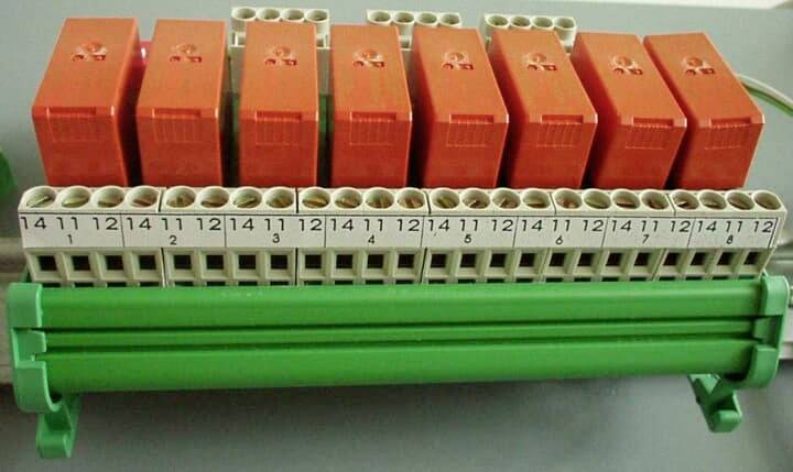 Eine Reihe von orangefarbenen Elektromodulen sitzt über einem beschrifteten Klemmenblock und einer grünen Montageschiene und zeigt eine industrielle Verdrahtungsbaugruppe.