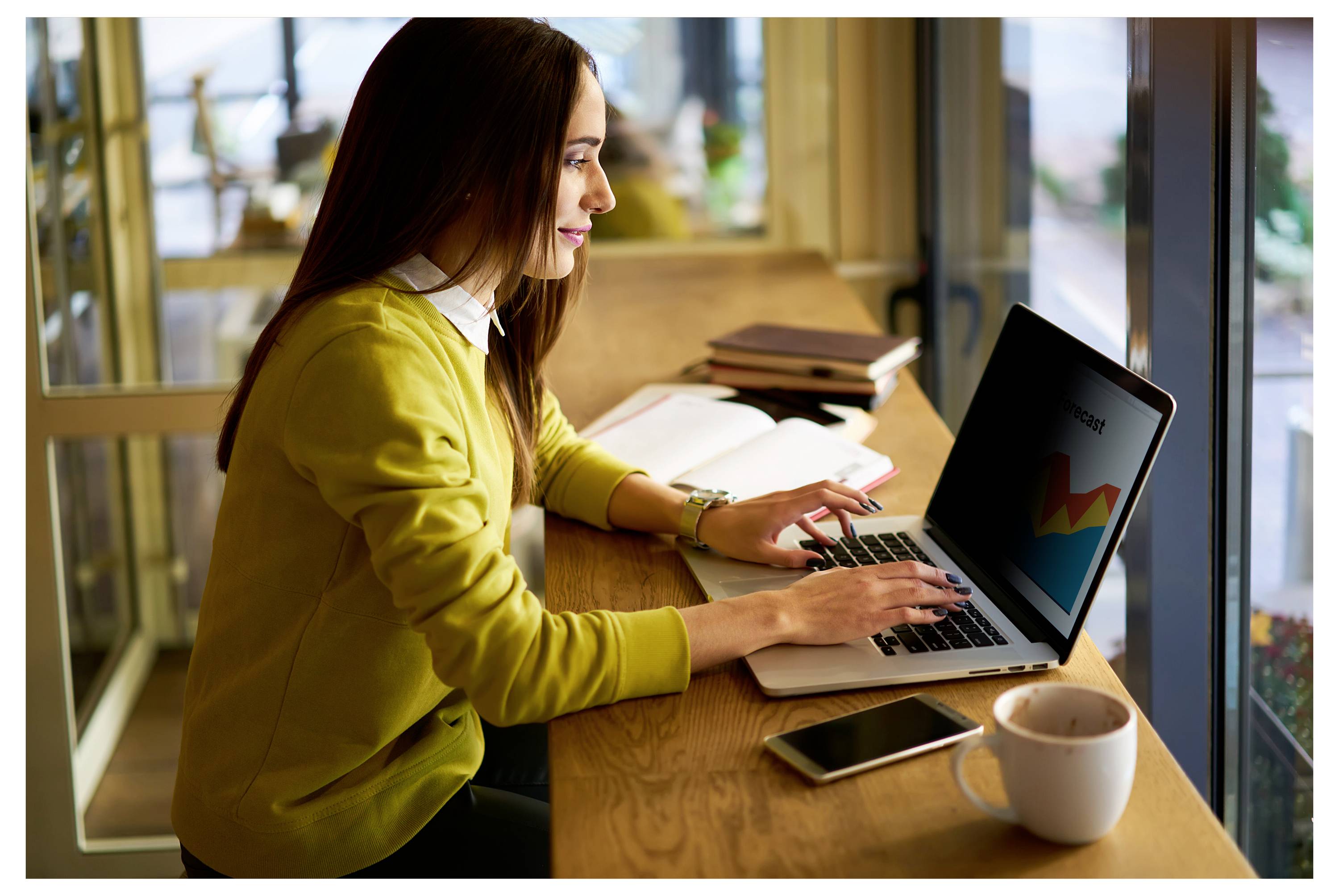 Frau, die an einem Laptopcomputer an einem Cafétisch neben einem Fenster arbeitet, mit einem Telefon, einer Kaffeetasse und einem offenen Notizbuch in der Nähe.