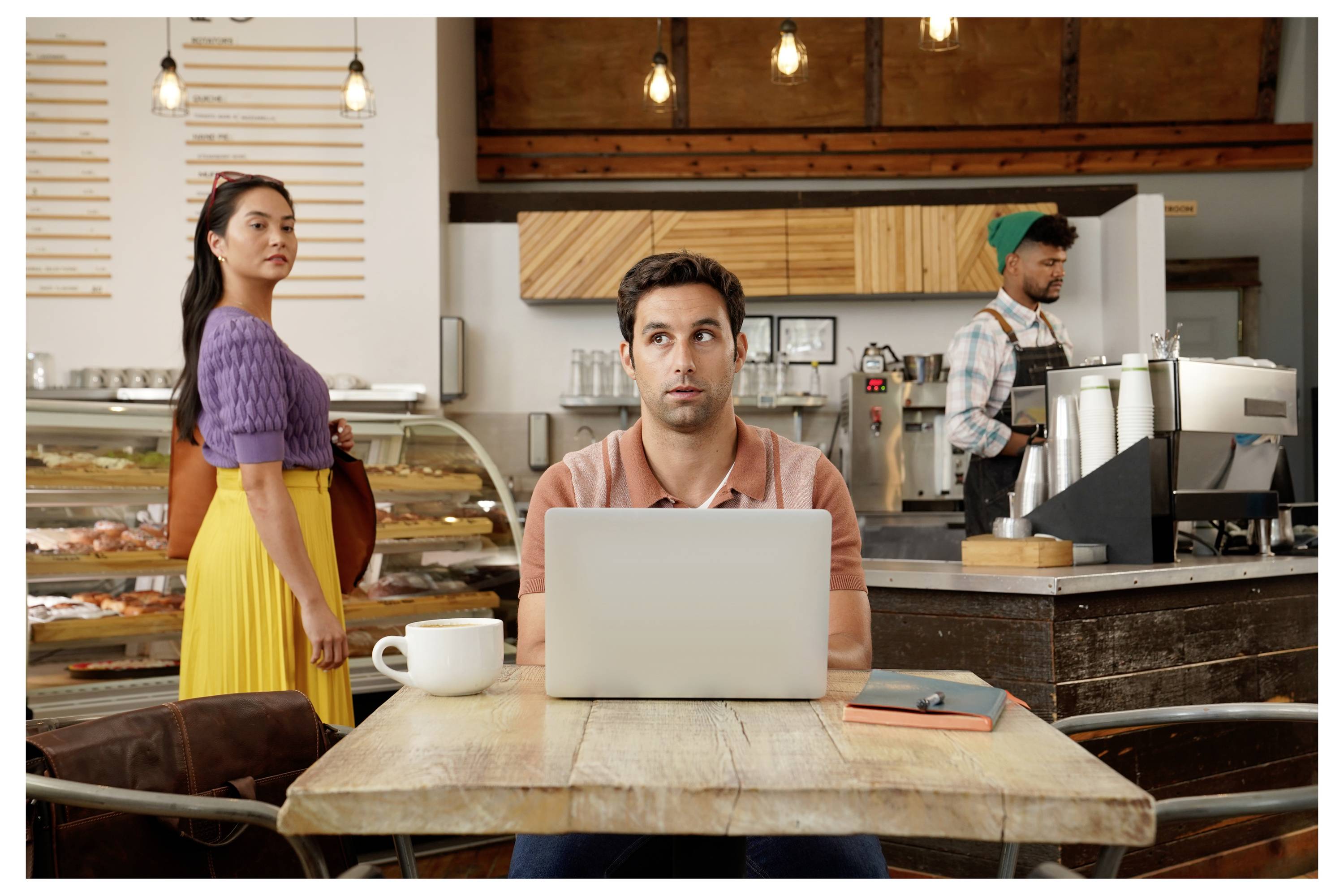 Ein Mann arbeitet an einem Laptop an einem Cafétisch, während eine Frau in der Nähe steht und ein Barista im Hintergrund Getränke zubereitet.