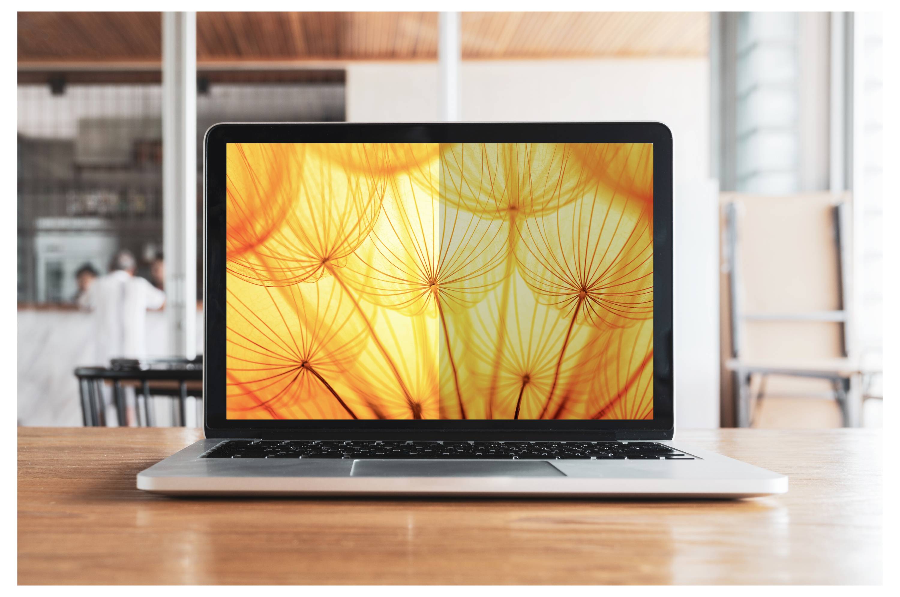 Laptop auf einem Holztisch geöffnet, das einen abstrakten orangefarbenen Blumenmuster auf einem hellen Bildschirm in einer hellen Büro- oder Café-Umgebung anzeigt.