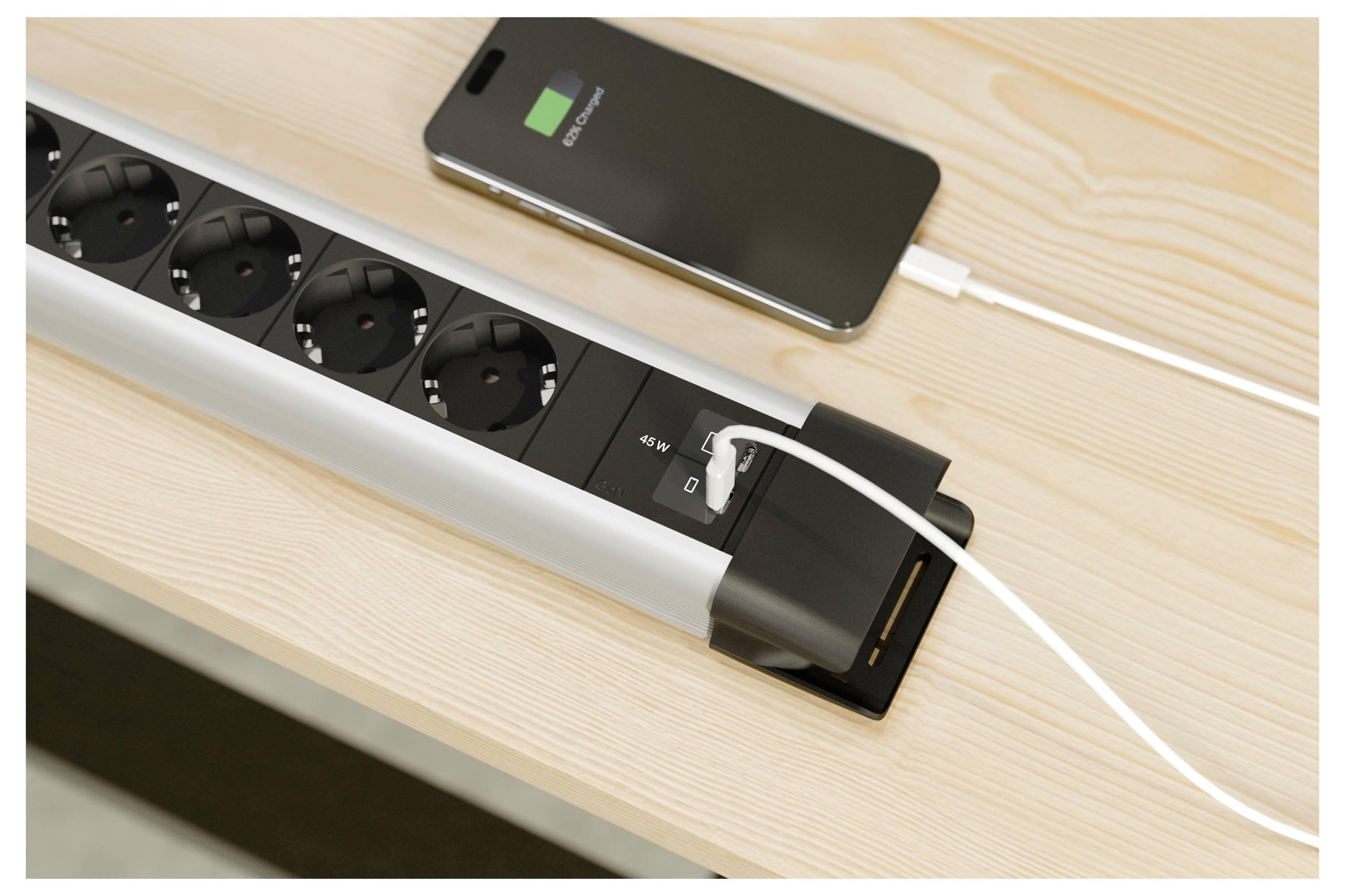 Smartphone wird von einer Steckdosenleiste mit einem USB-Kabel auf einem Holzschreibtisch aufgeladen, wobei eine angeschlossene Steckdose und der Ladestatus angezeigt werden.