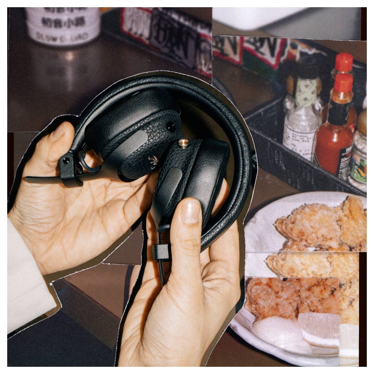 Hände halten schwarze Over-Ear-Kopfhörer über einen Tisch mit Hähnchen, Würzmitteln und einem Glas; lockere Essensatmosphäre.