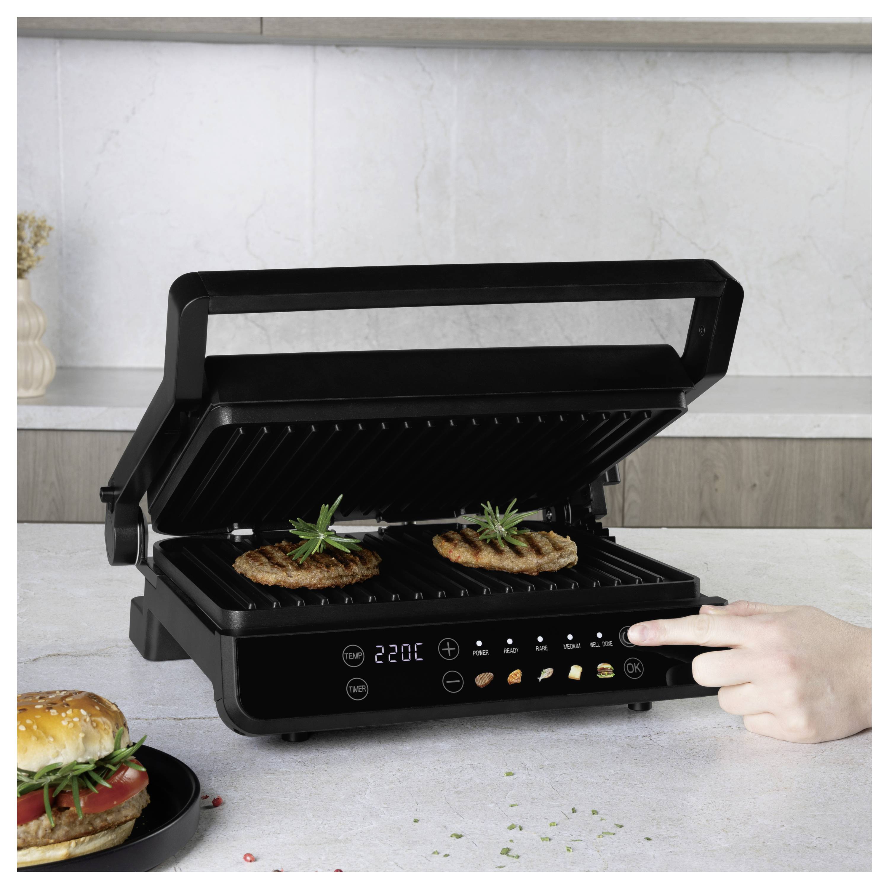 Ein tragbarer elektrischer Grill auf der Arbeitsplatte ist offen und bereitet zwei Patties zu; eine Hand drückt auf das Bedienfeld, während ein Burger auf einem Teller in der Nähe liegt.