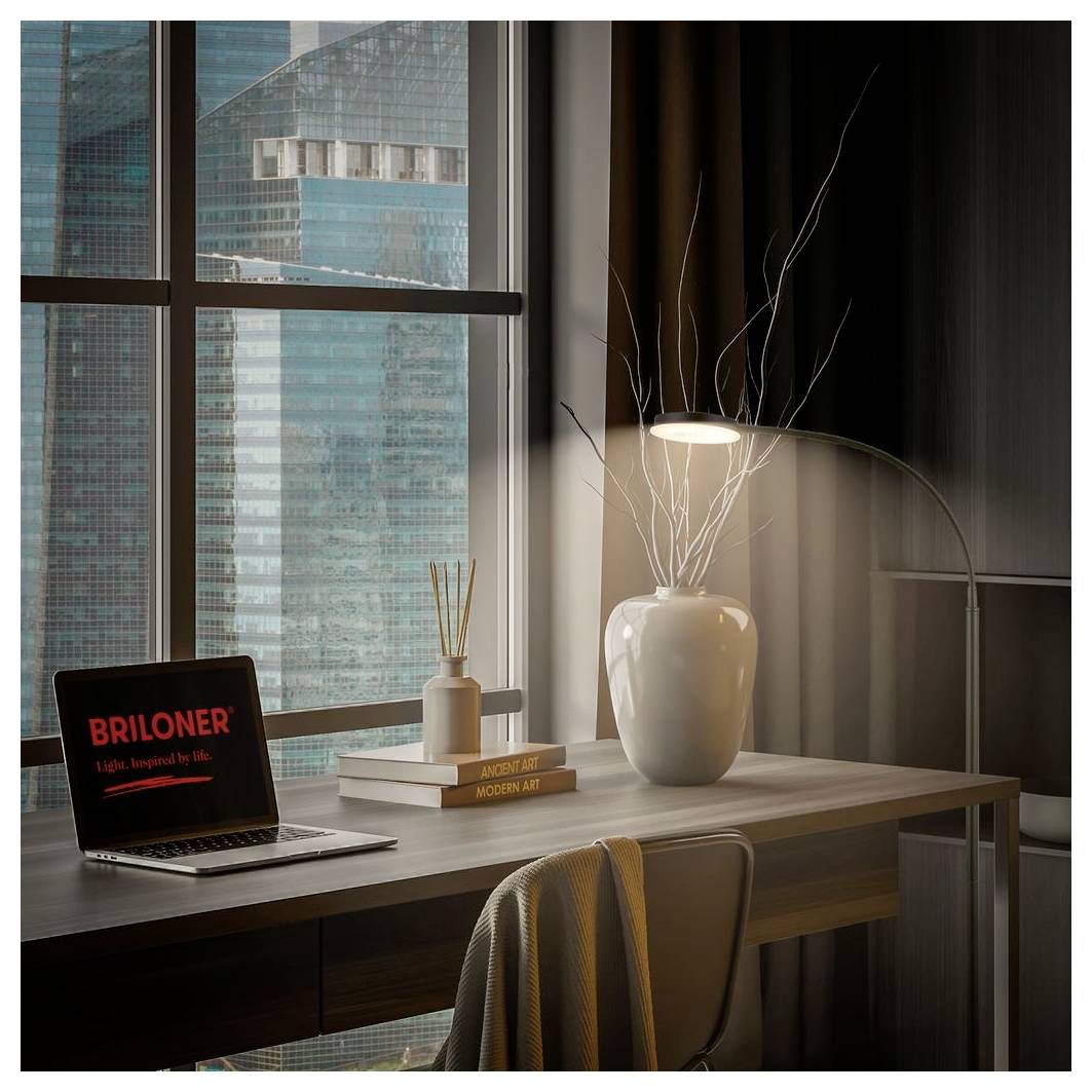 Moderner Homeoffice-Schreibtisch am Fenster mit einem Laptop, gestapelten Büchern, einer Vasenlamp und Blick auf Hochhäuser der Stadt draußen.