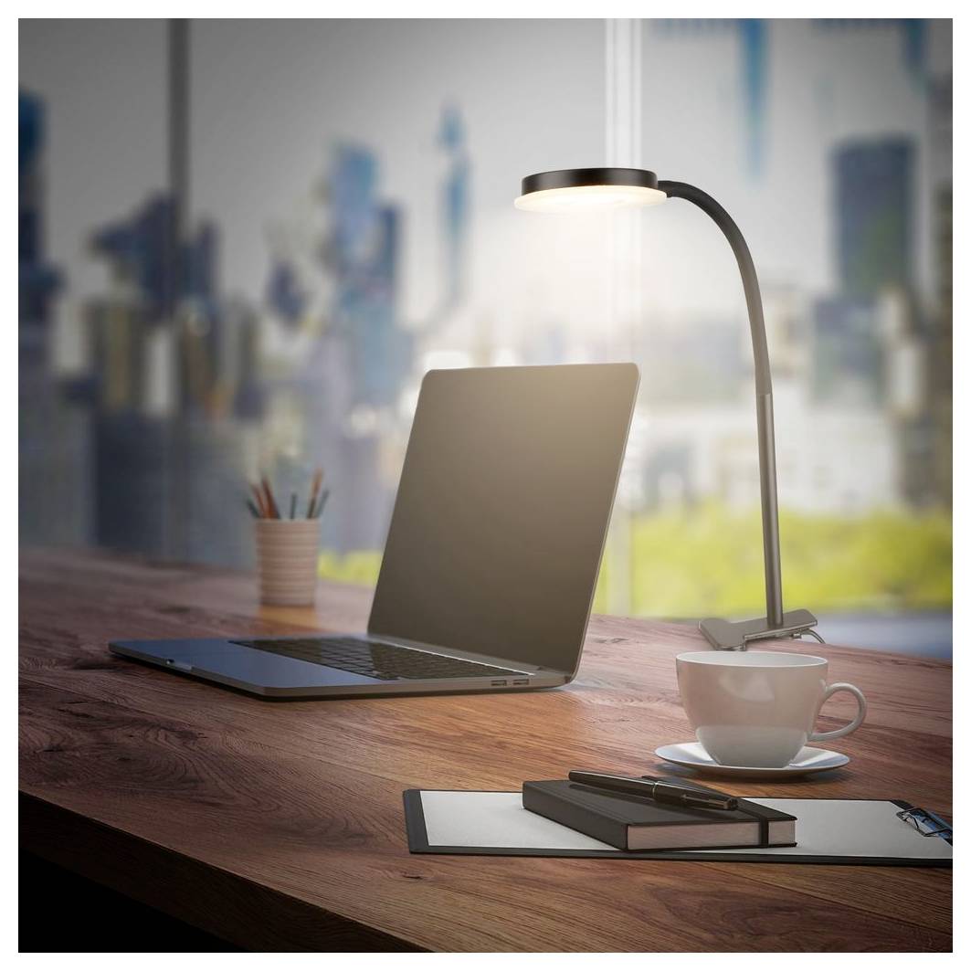 Ein Laptop auf einem Holzschreibtisch neben einer brennenden Lampe, einer Kaffeetasse, Notizbüchern und einem Stifthalter, mit einer unscharfen Stadtsilhouette im Hintergrund.