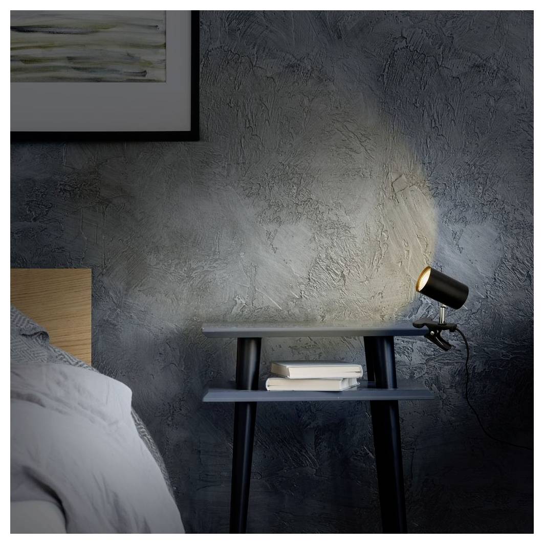 Eine Schlafzimmerecke mit einem Bett, einem kleinen Nachttisch, gestapelten Büchern und einer Strahlerleuchte, die Licht auf eine strukturierte graue Wand wirft.