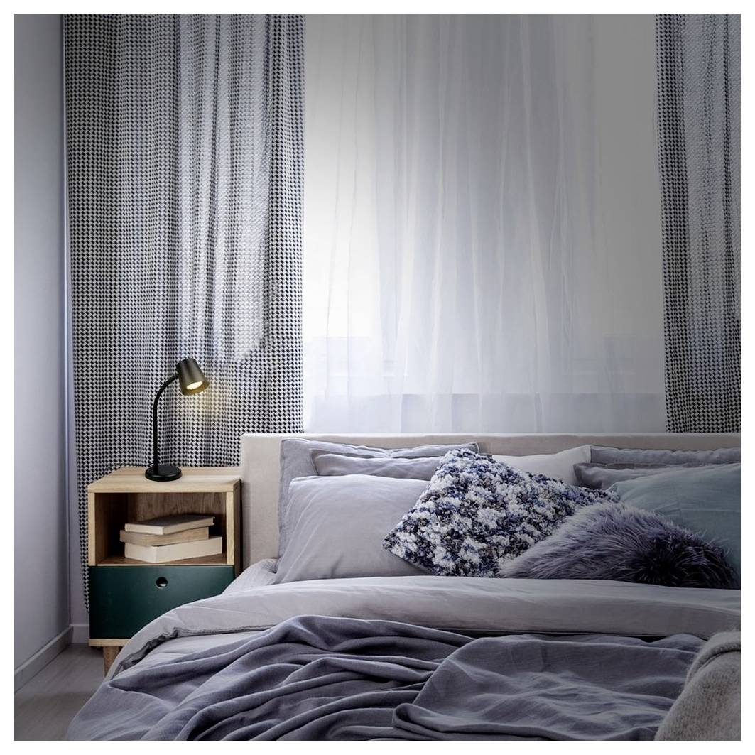 Ein ordentlich gemachtes Bett in einem sanft beleuchteten Schlafzimmer neben einem hölzernen Nachttisch mit einer Lampe und Büchern, mit transparenten Vorhängen über einem Fenster.