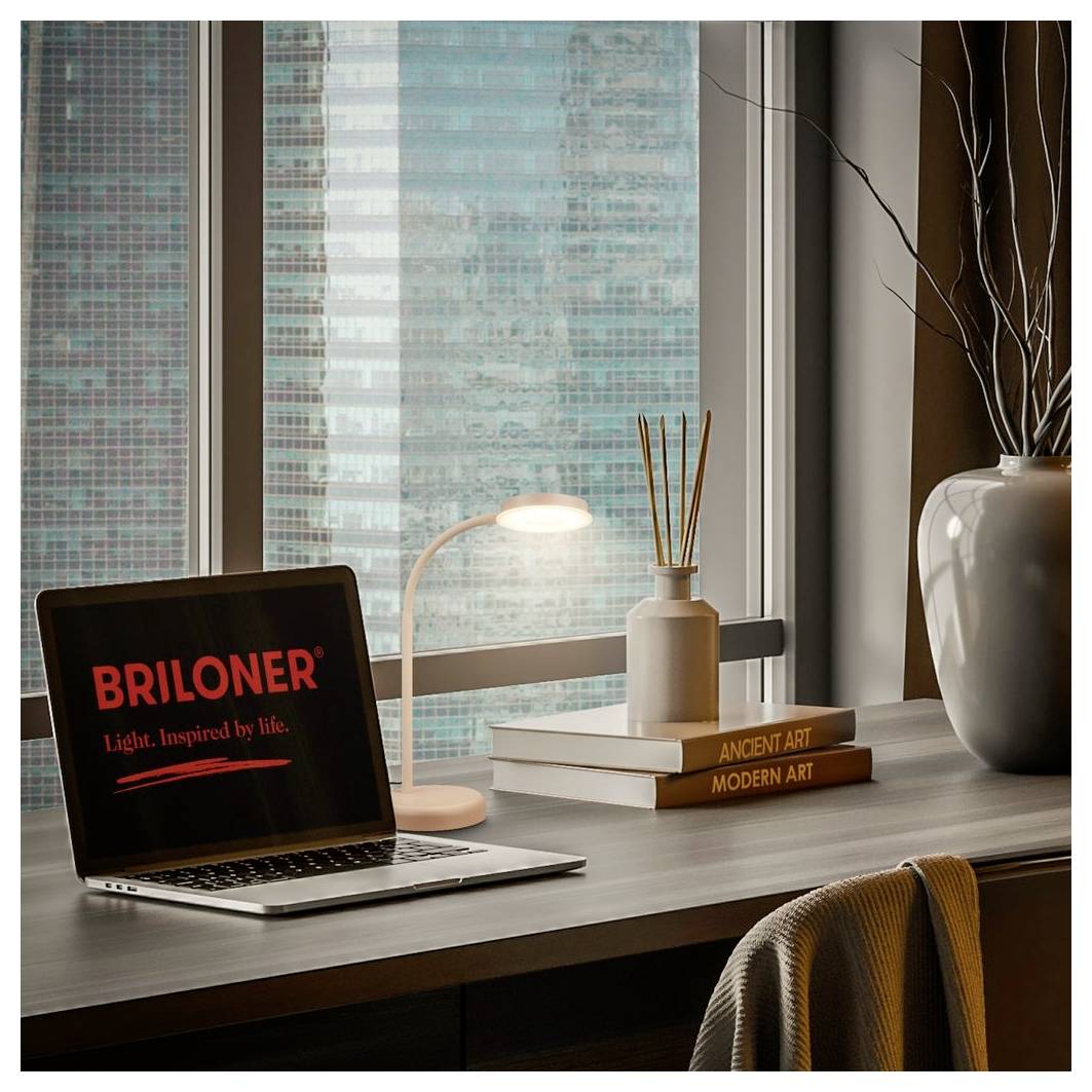 Laptop auf dem Schreibtisch mit der Anzeige "BRILONER" neben einer leuchtenden Lampe, Büchern und einer Vase bei einem Fenster mit Stadtgebäuden draußen.